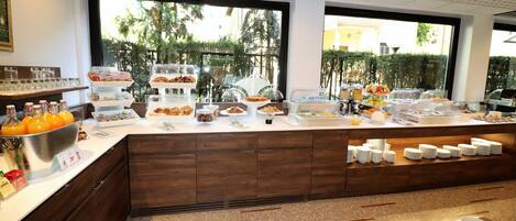 A buffet area with various food items and beverages, a large window with a view of greenery, and a modern kitchen in the background.