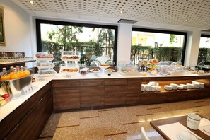 En buffet med forskellige retter og drikkevarer, et stort vindue med udsigt over grønne områder, og et moderne køkken i baggrunden.