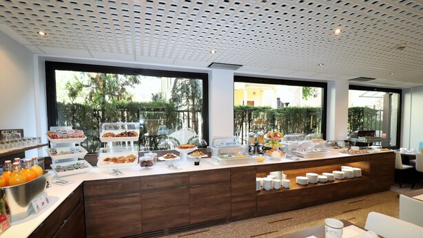 Un bufé de desayuno con diversas opciones de comida y bebidas, un área de estar con mesas y ventanas amplias que ofrecen vistas a zonas verdes.