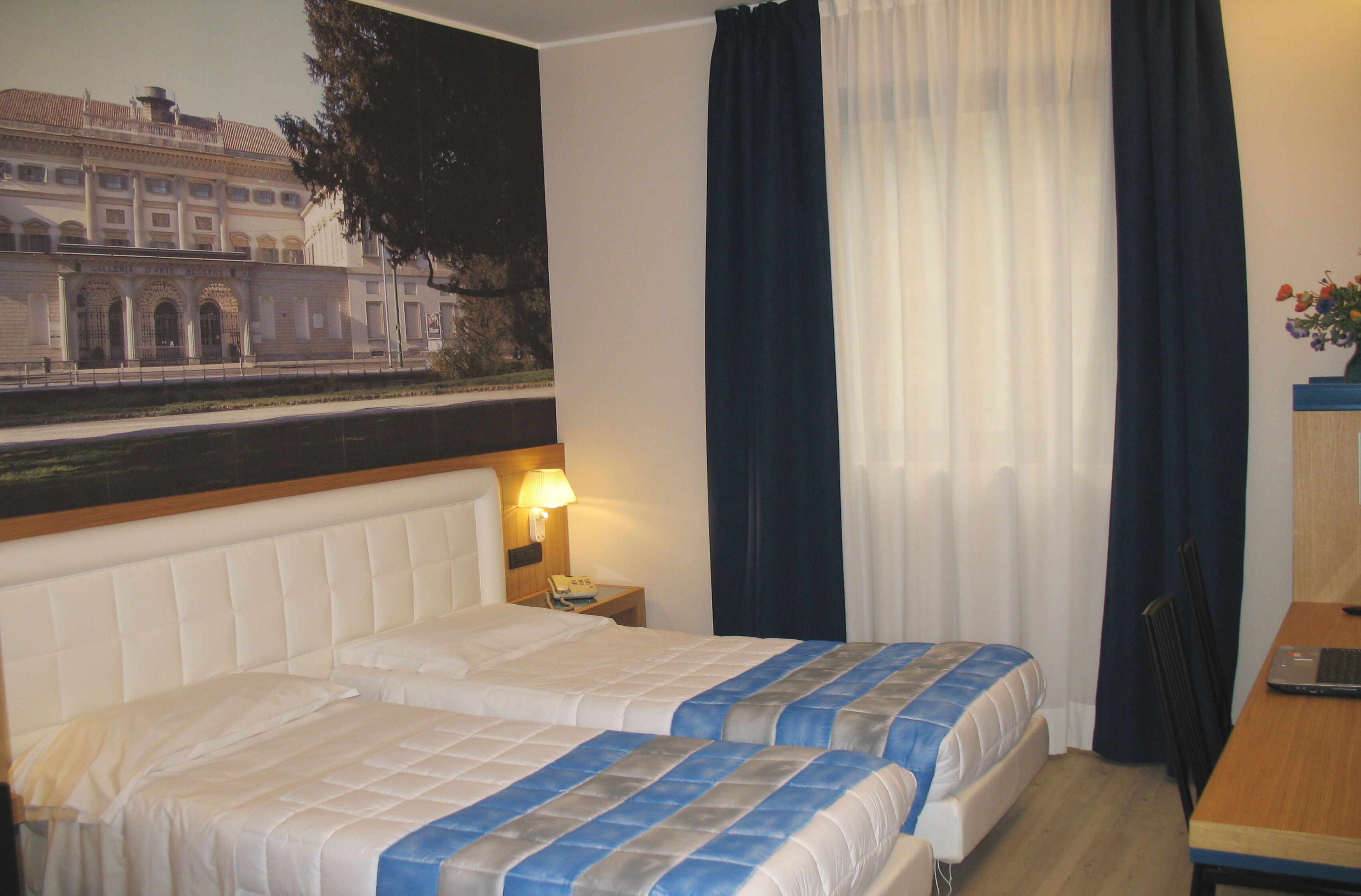 Habitación de hotel con dos camas, un escritorio, una silla, una lámpara y un cuadro de un edificio en la pared.