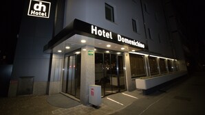 Entrada de hotel con un letrero que dice "Hotel Domanichine", con un diseño moderno que incluye puertas de vidrio y una fachada bien iluminada.