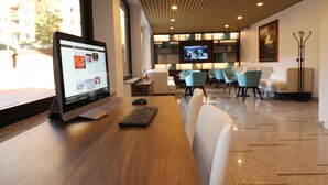Un moderno vestíbulo de hotel con un mostrador de recepción de madera, un monitor de computadora y una pantalla de televisión que muestra un noticiero. Hay varias sillas y un sofá en la zona de estar.