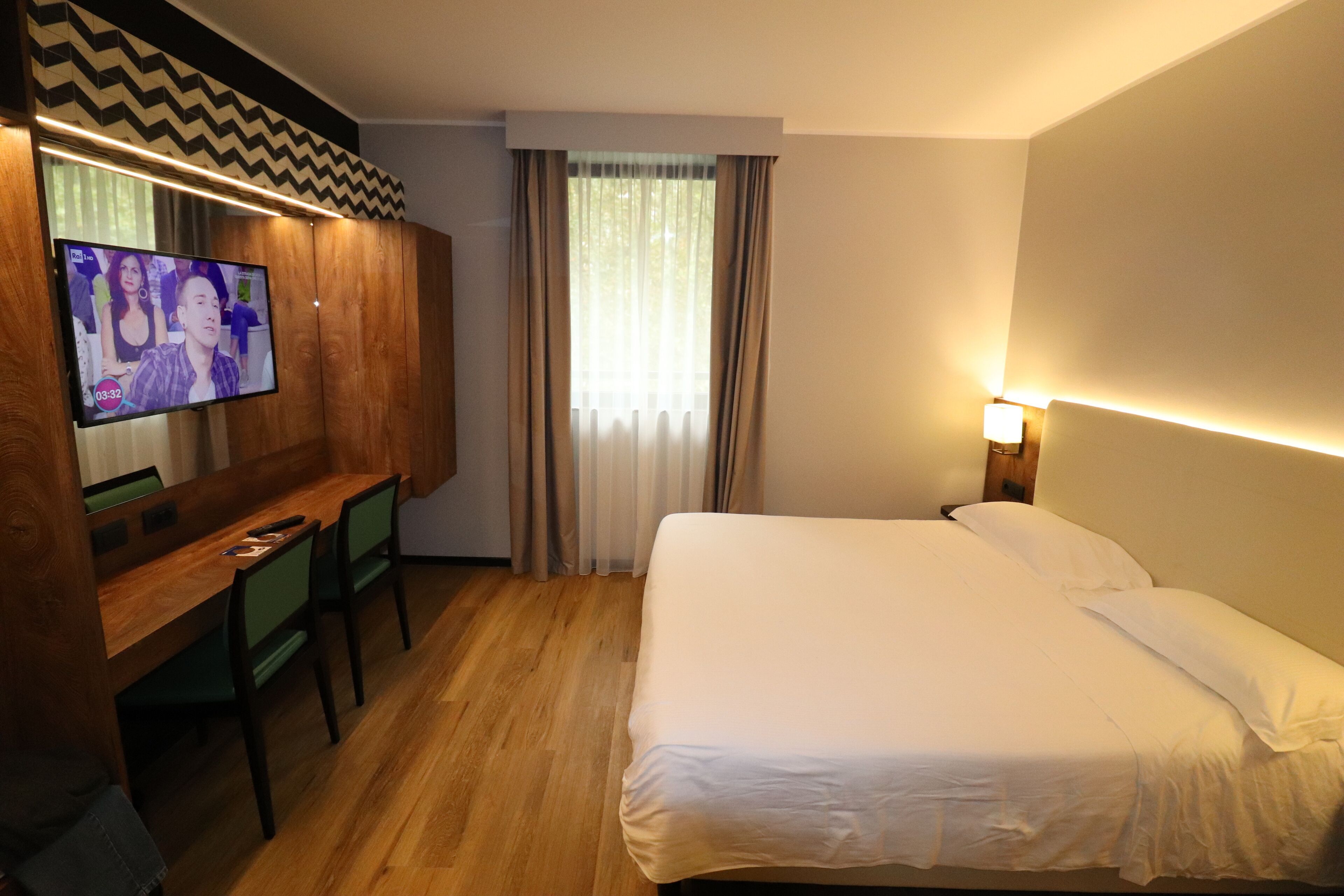 A hotel room with a bed, a desk with a chair, a television, and a window with curtains.