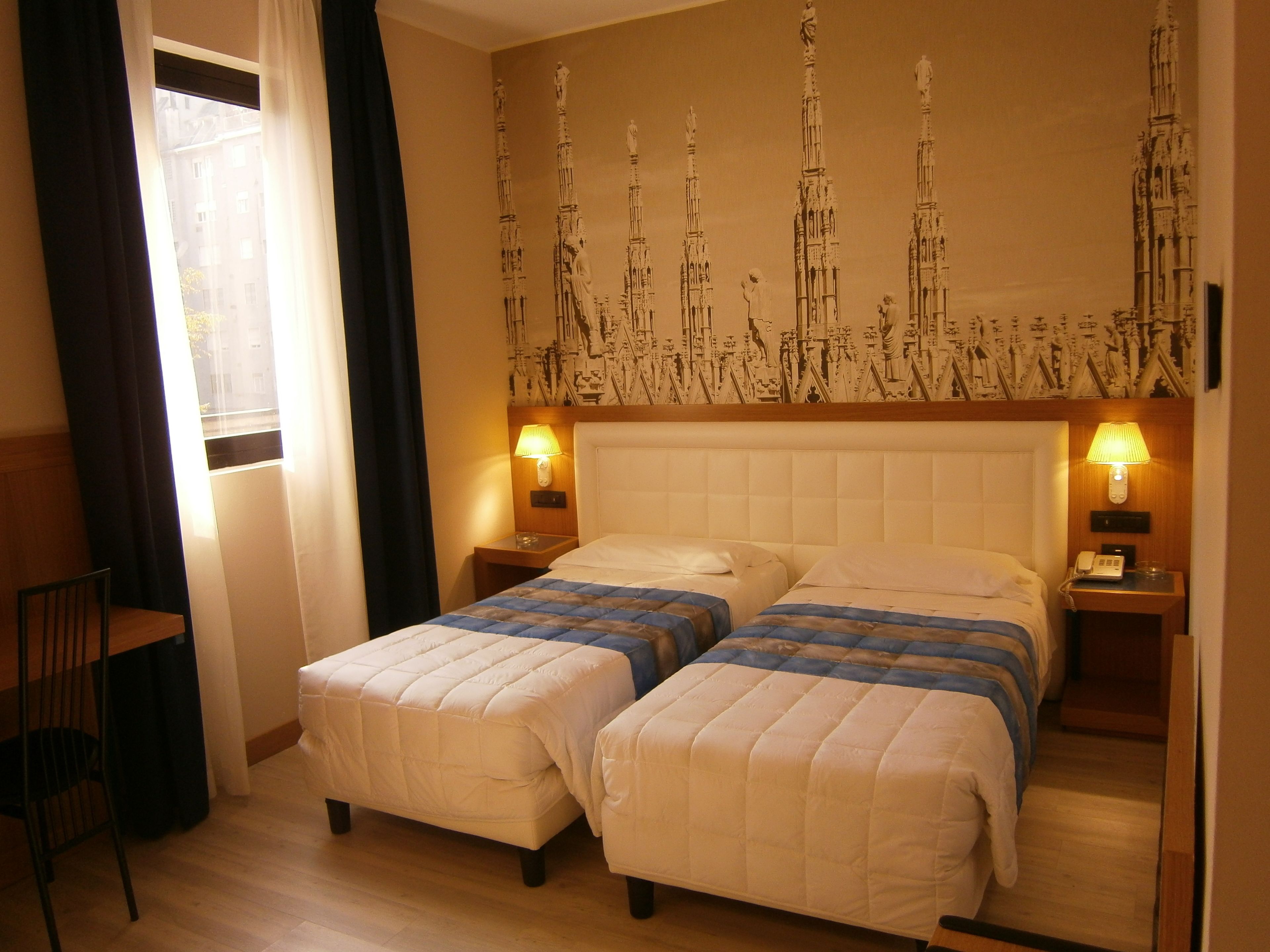 A hotel room with two beds, a chair, a nightstand with a lamp, and a wall with a patterned design.