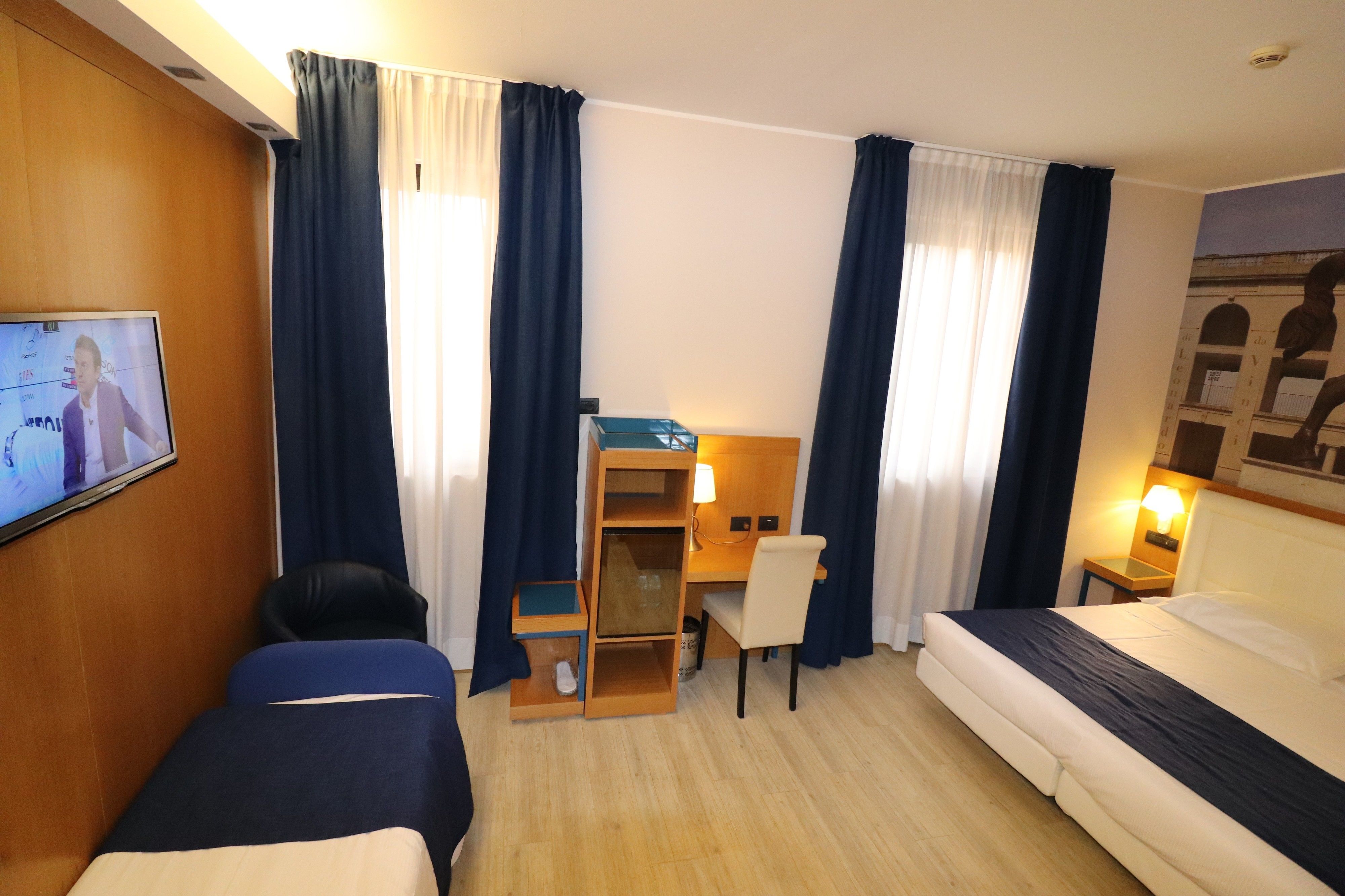 A hotel room with two beds, a desk, a chair, a TV, and a lamp.