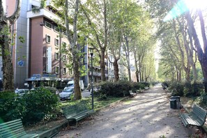 En allé med træer, bænke, et hotel og parkerede biler.