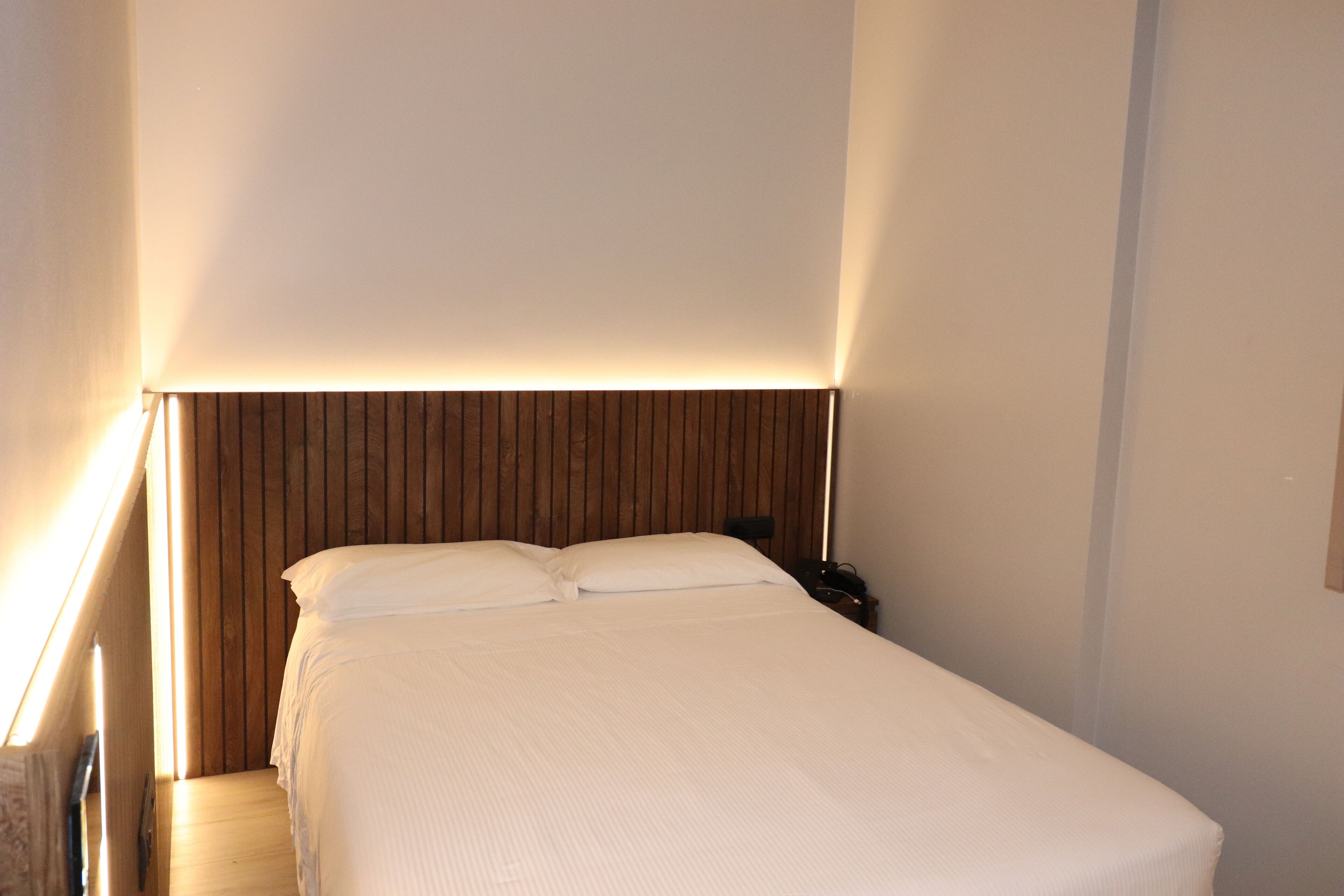 Una cama bien tendida en una habitación de hotel, con cabecera de madera y una lámpara de noche.