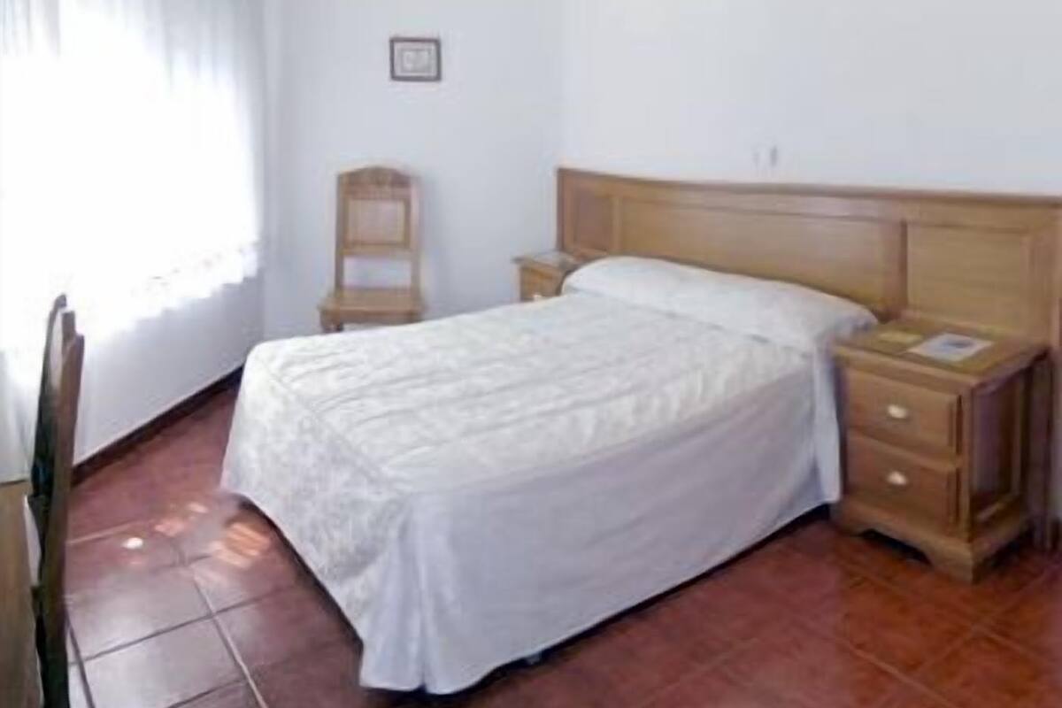 Une chambre avec un lit simple, une table de chevet en bois, une chaise et une fenêtre avec des rideaux.