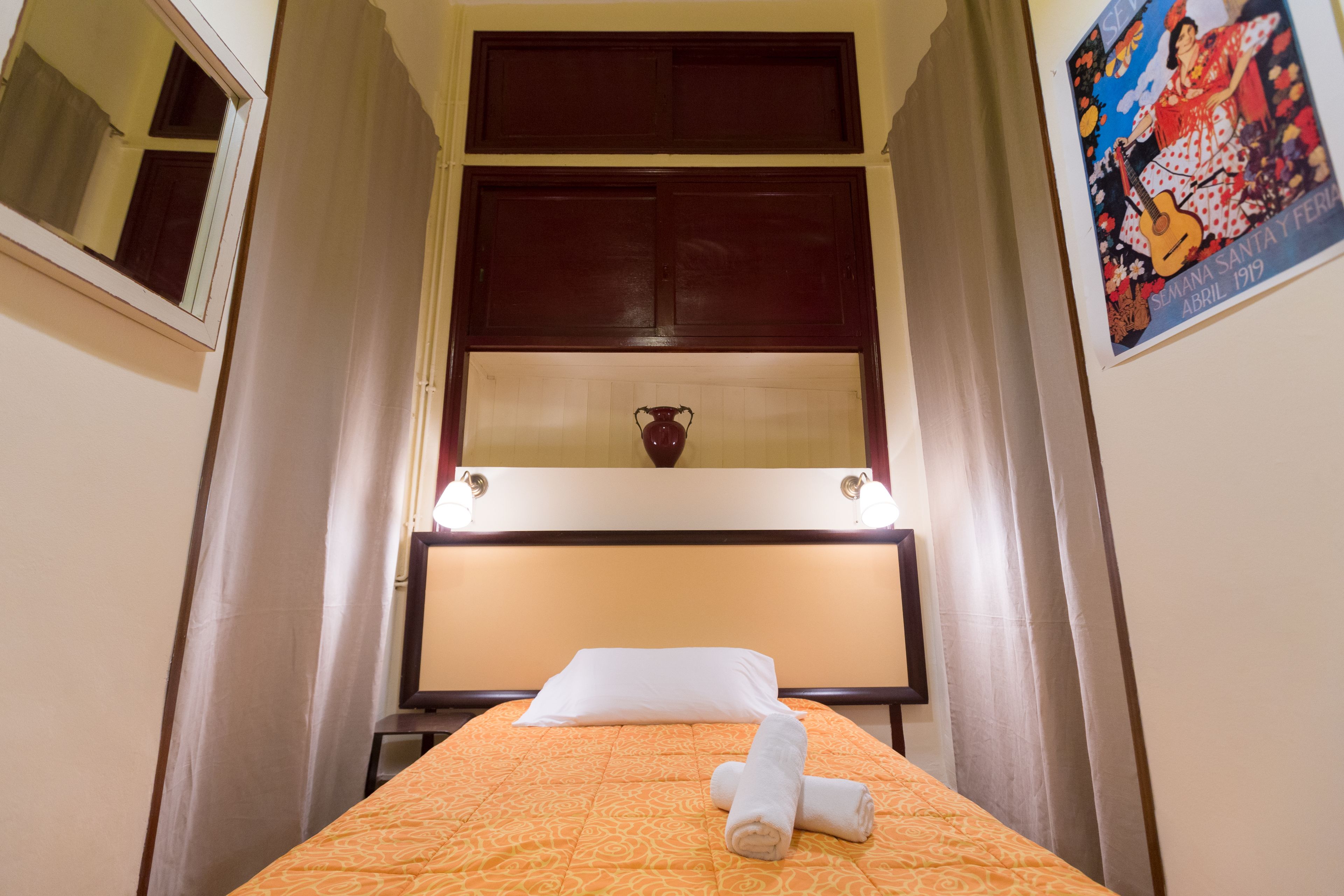 Une chambre d’hôtel avec un lit, une table de chevet, un miroir, une fenêtre avec des rideaux et une affiche accrochée au mur.