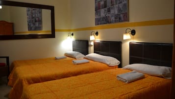 A hotel room with two beds, each with an orange patterned bedspread, white pillows, and neatly folded towels. There are wall-mounted lamps above each bed, and a large mirror on the left wall.