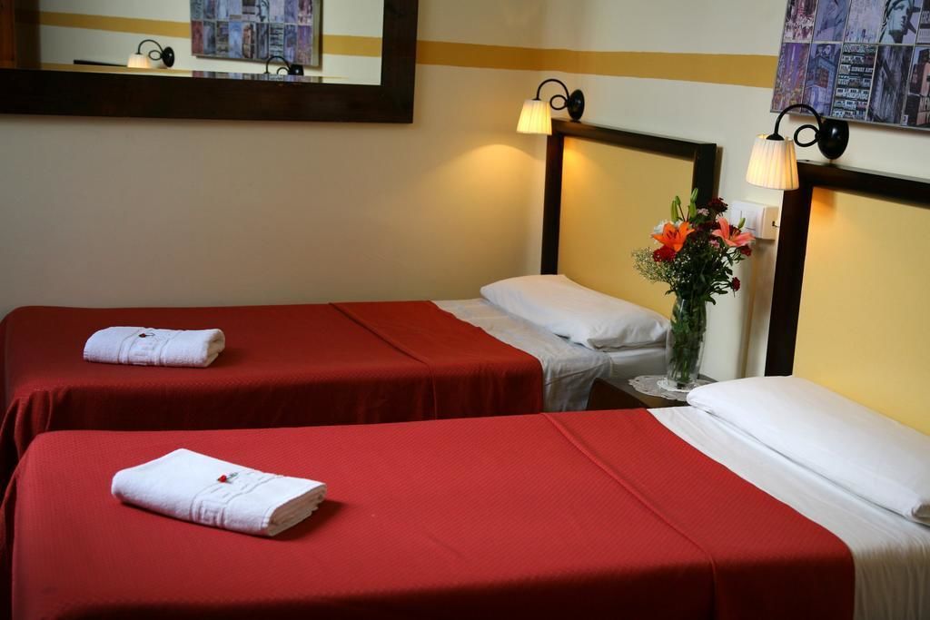Een hotelkamer met twee enkele bedden, elk met een rode sprei en witte kussens. Daartussen staat een klein tafeltje met een bloemenvaas.