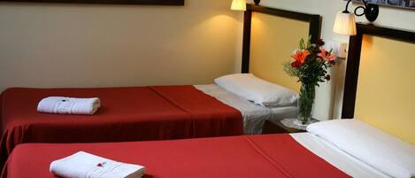 Een hotelkamer met twee enkele bedden, elk met een rode sprei en witte kussens. Daartussen staat een klein tafeltje met een bloemenvaas.