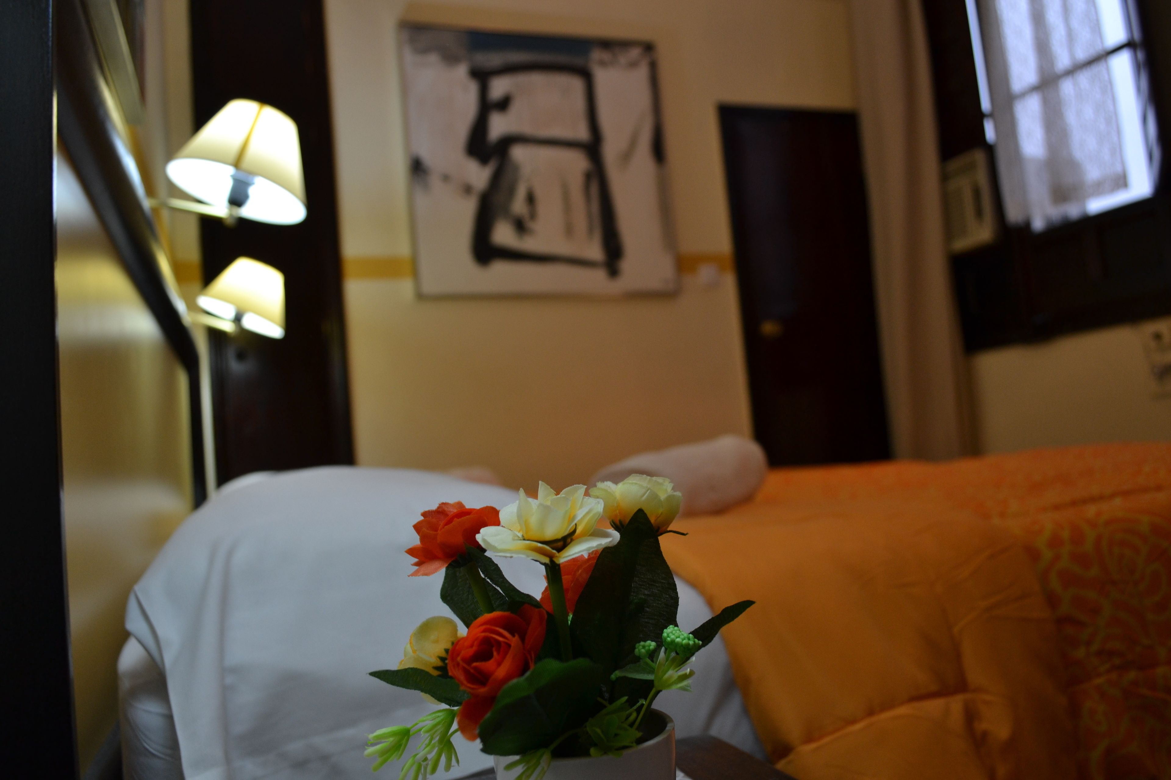 Una habitación de hotel con una cama, una mesita de noche con un florero, una lámpara y una obra de arte en la pared.