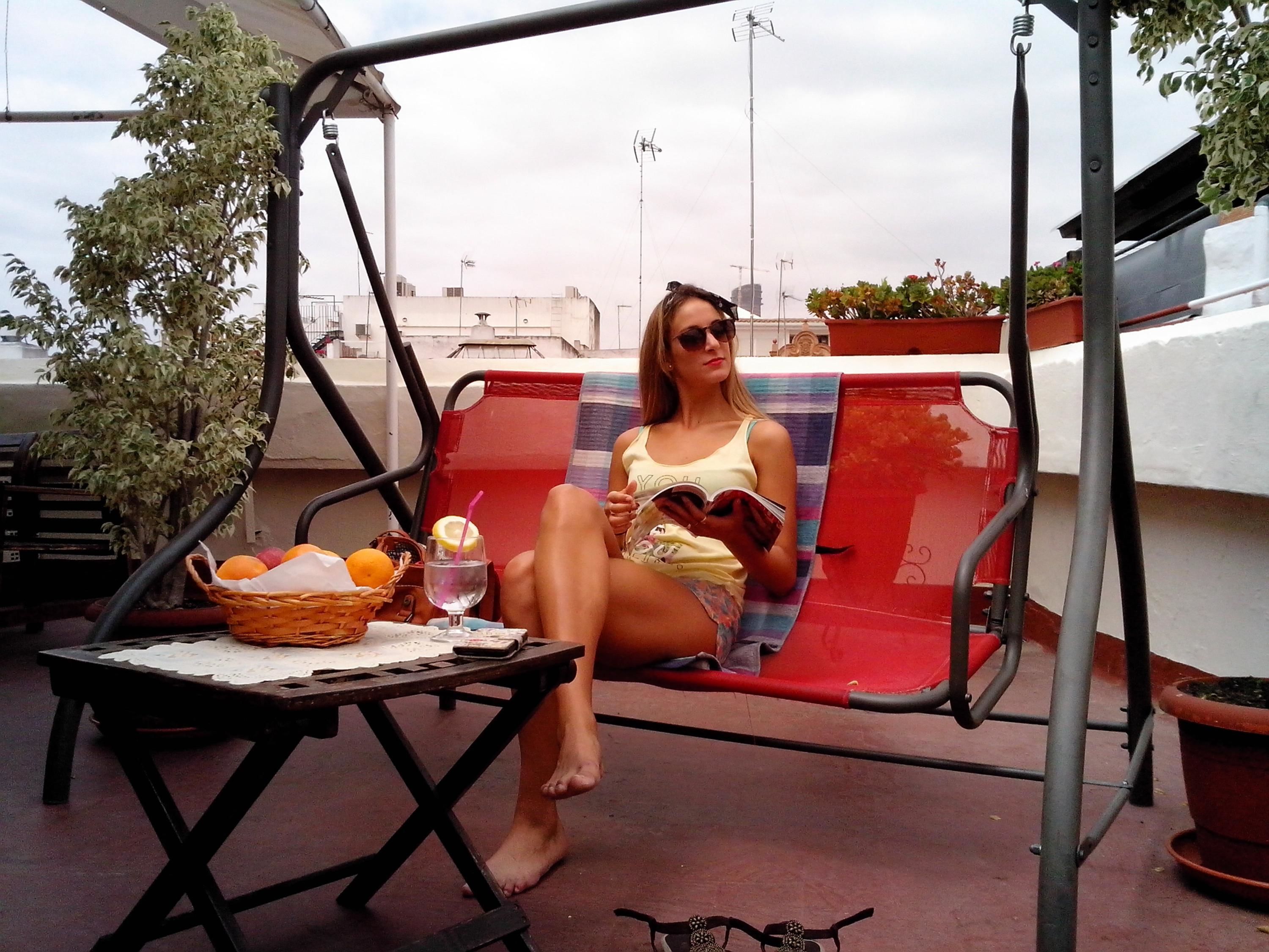 A person sitting on a red and black swing chair, reading a book. There's a table with a basket of fruits and a glass of water with a lemon.