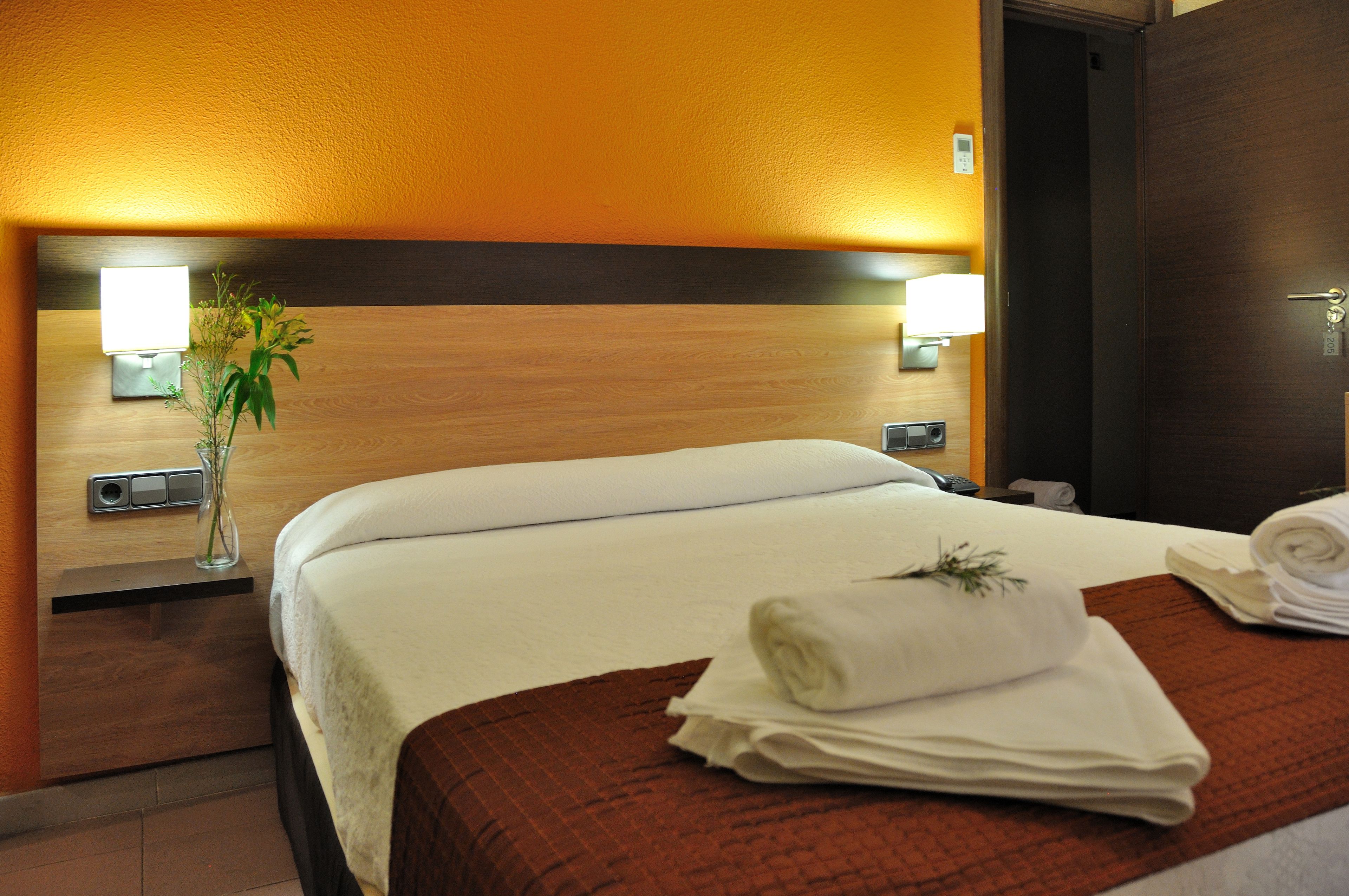 A hotel room with a neatly made bed, a nightstand with a lamp, and a wall-mounted light fixture.