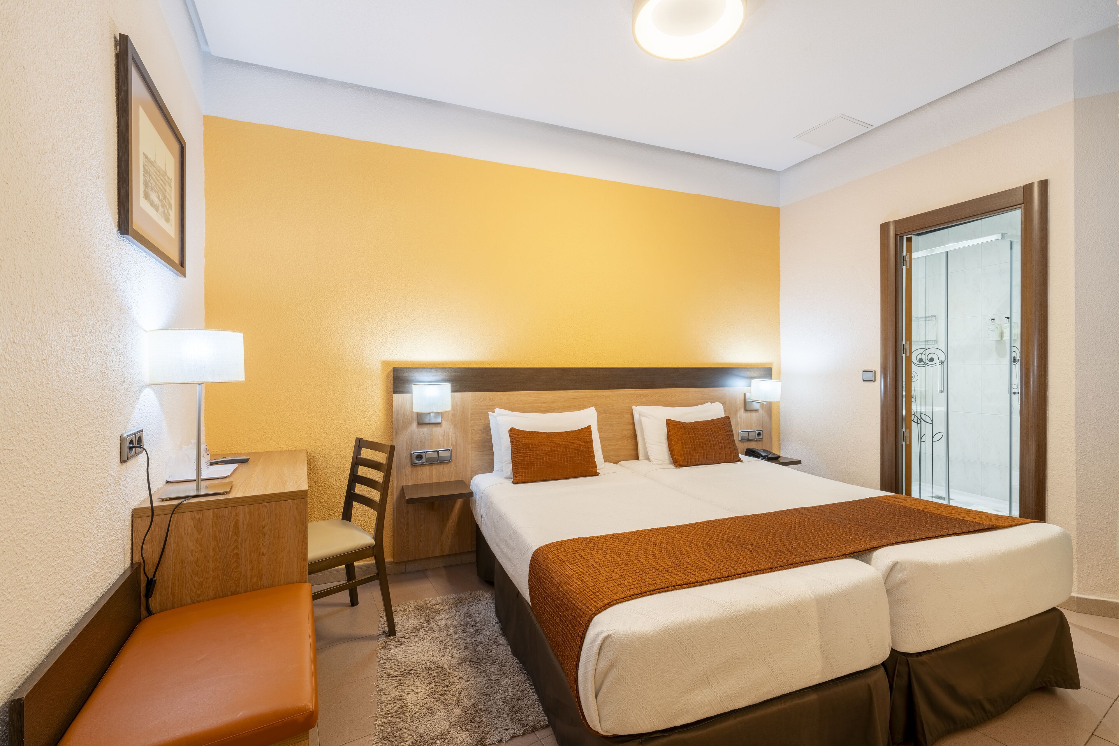 A hotel room with a large bed, a desk with a chair, a bench, and a bathroom with a glass door.