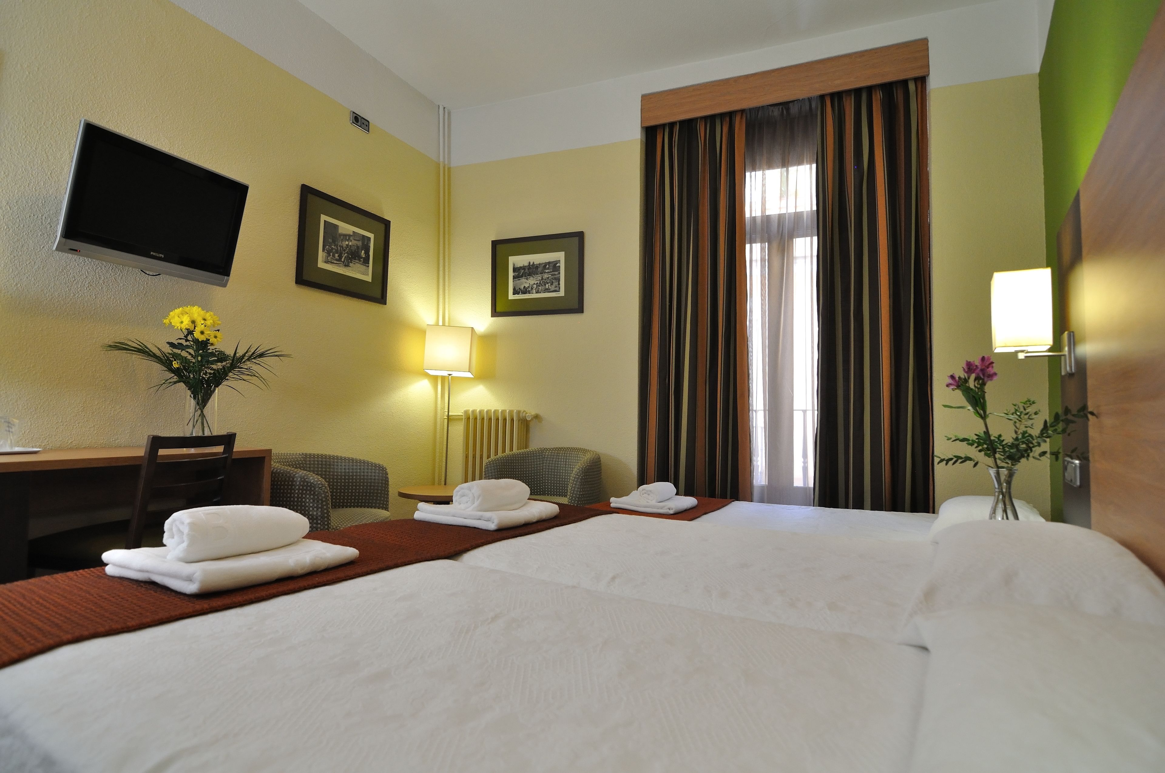 A hotel room with a bed, a desk, a chair, a television, and framed pictures on the wall.