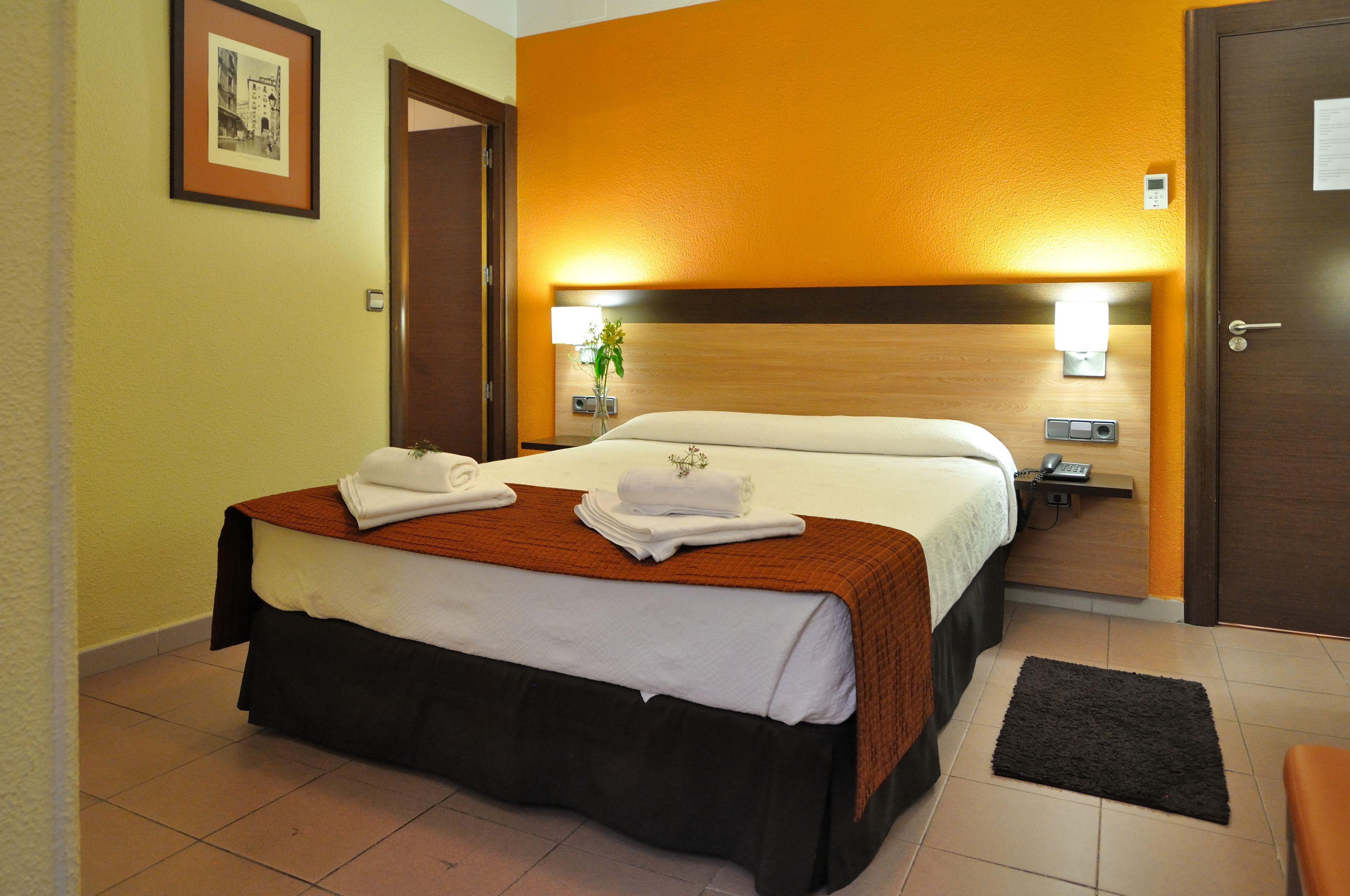 A hotel room with a bed, a nightstand, a wall-mounted lamp, and a door.