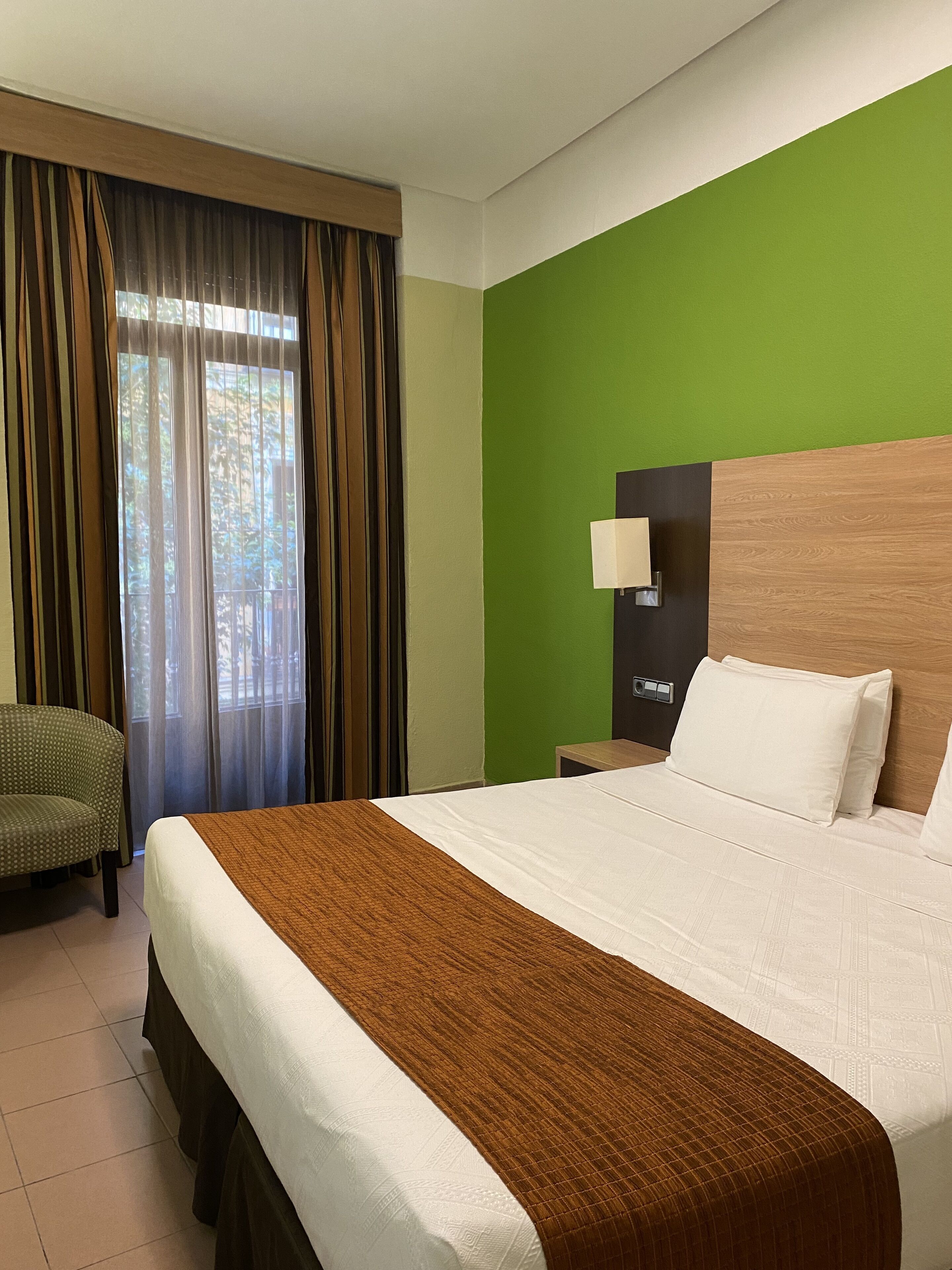A hotel room with a large bed, a green accent wall, a window with curtains, a nightstand with a lamp, and a chair.