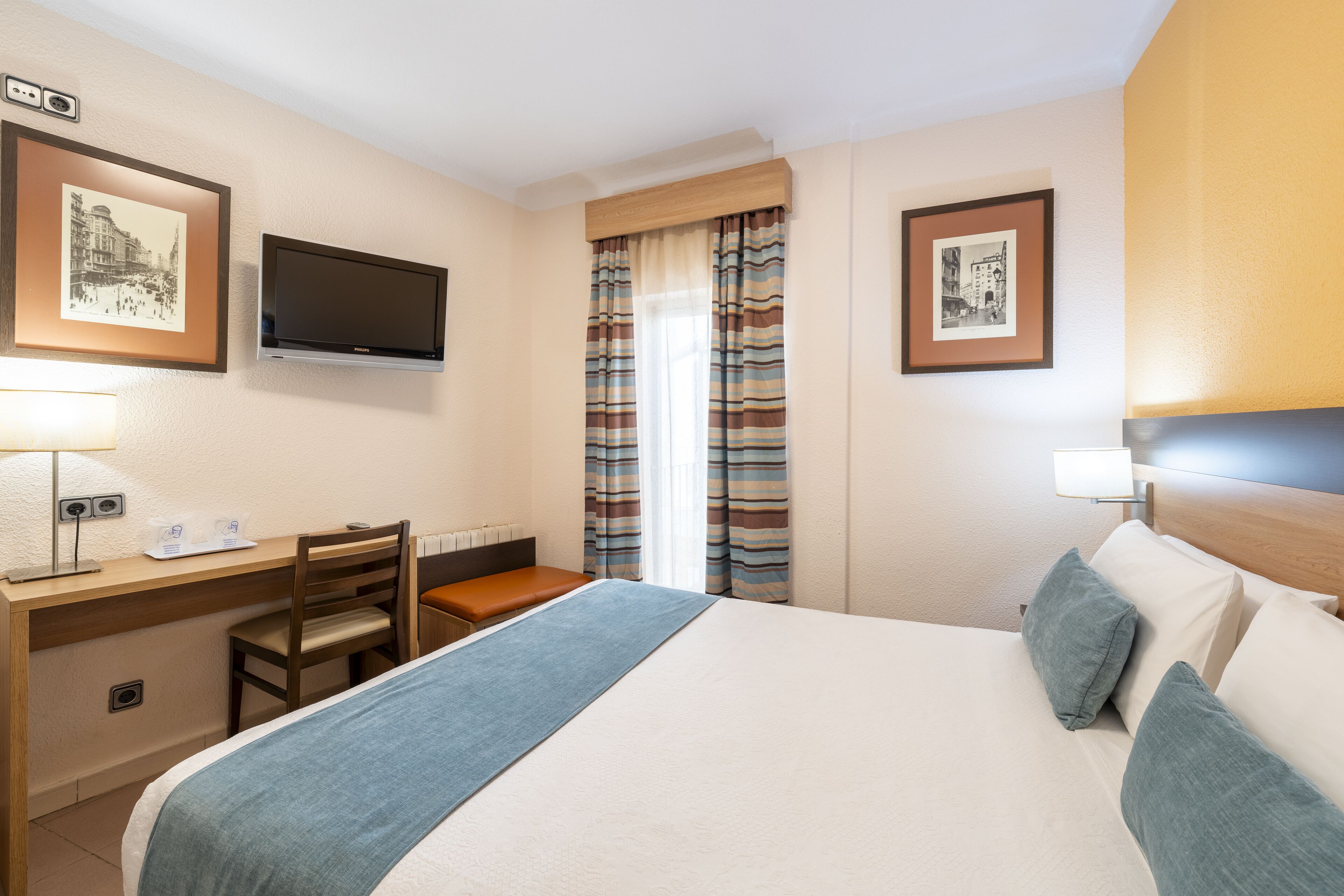 Ett hotellrum med en säng, ett skrivbord, en stol, en tv och inramade tavlor på väggarna.