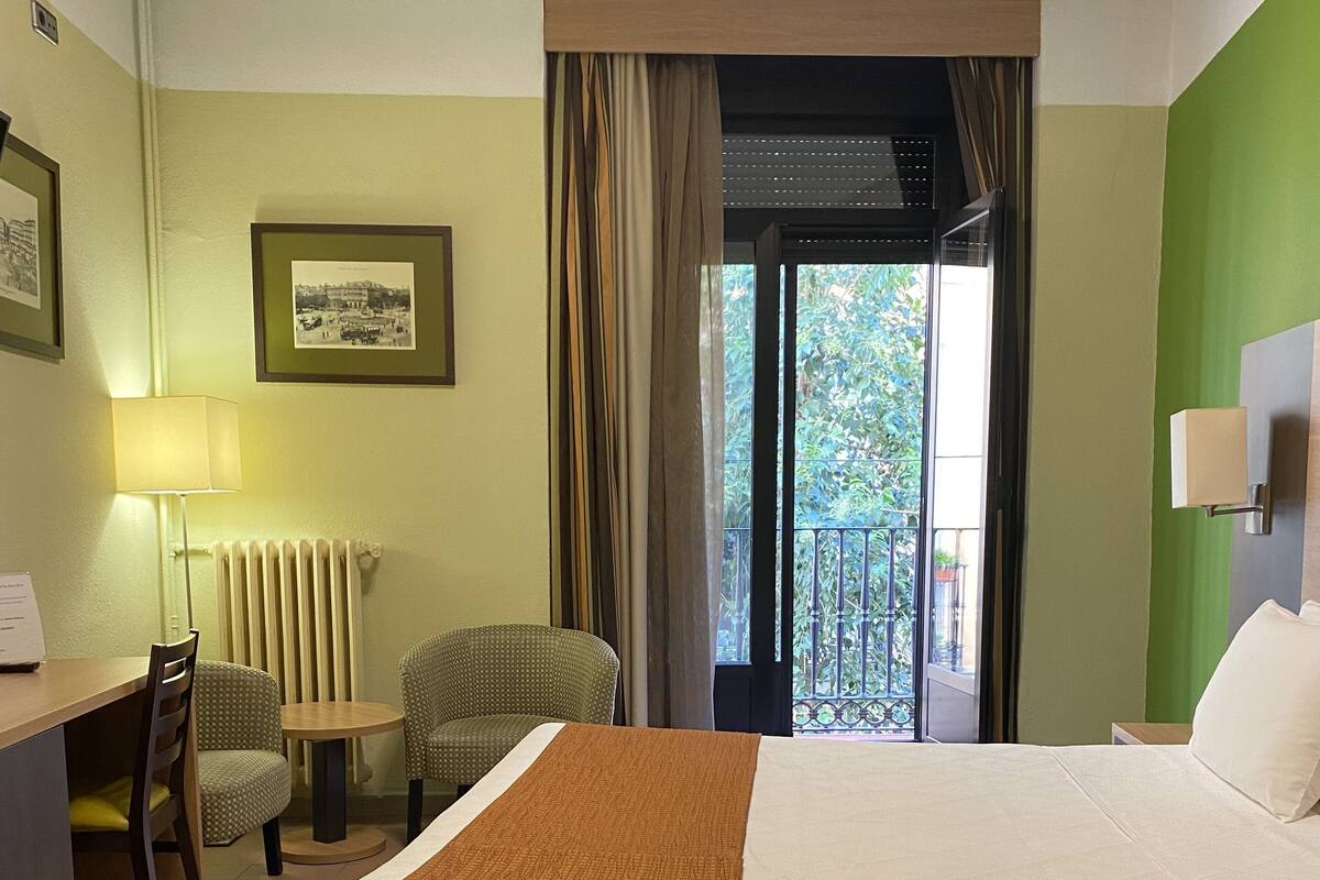 A hotel room with a bed, a desk, a chair, a lamp, a framed picture on the wall, and a view of the outdoors through a window.