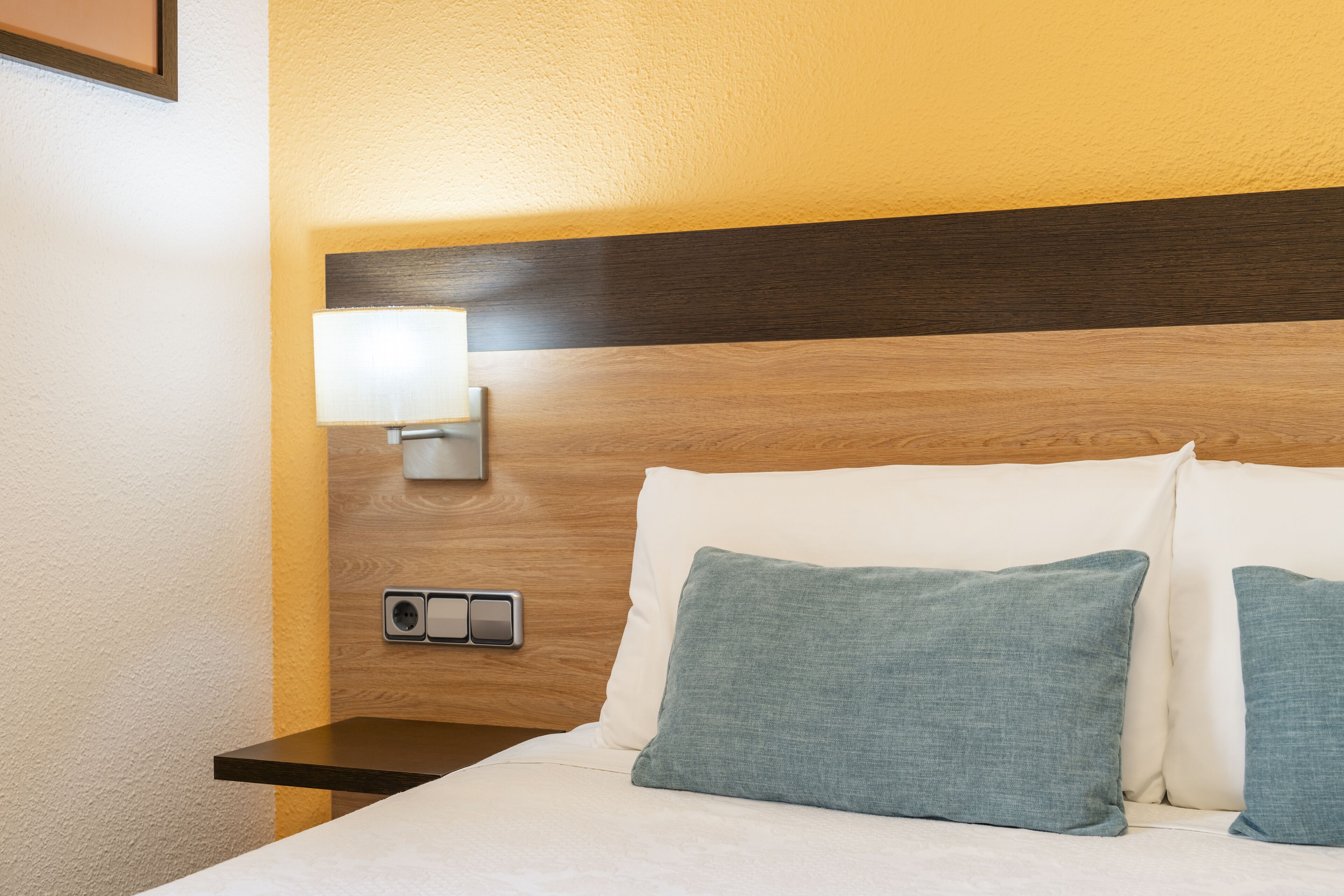 Ein Hotelzimmer mit einem Holzkopfteil, einer Nachttischlampe, einer Steckdose und zwei Kissen.