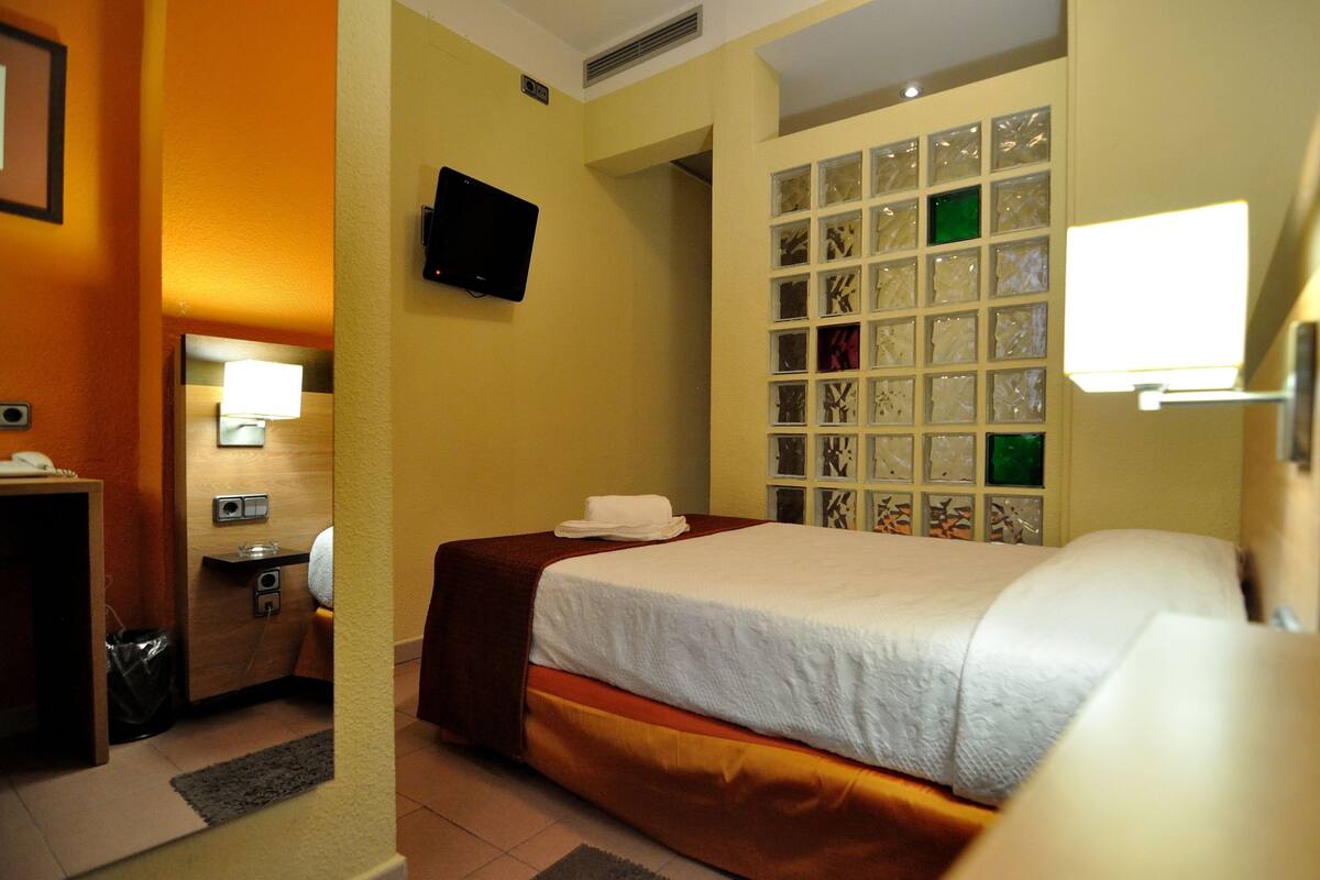 A hotel room with a bed, a desk with a lamp, a TV, and a window with decorative glass panels.