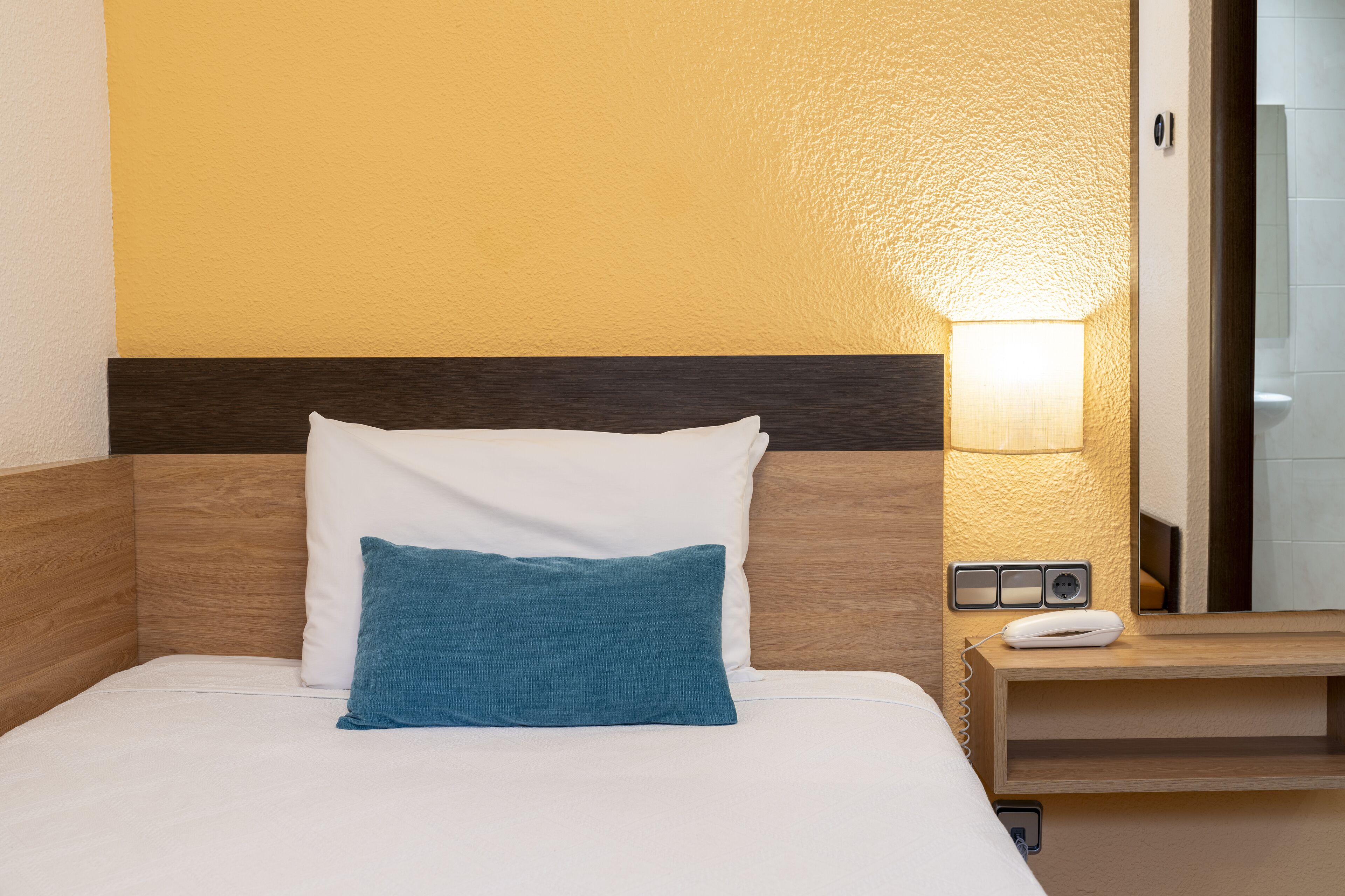 A hotel room with a bed, a nightstand, a lamp, and a power outlet.