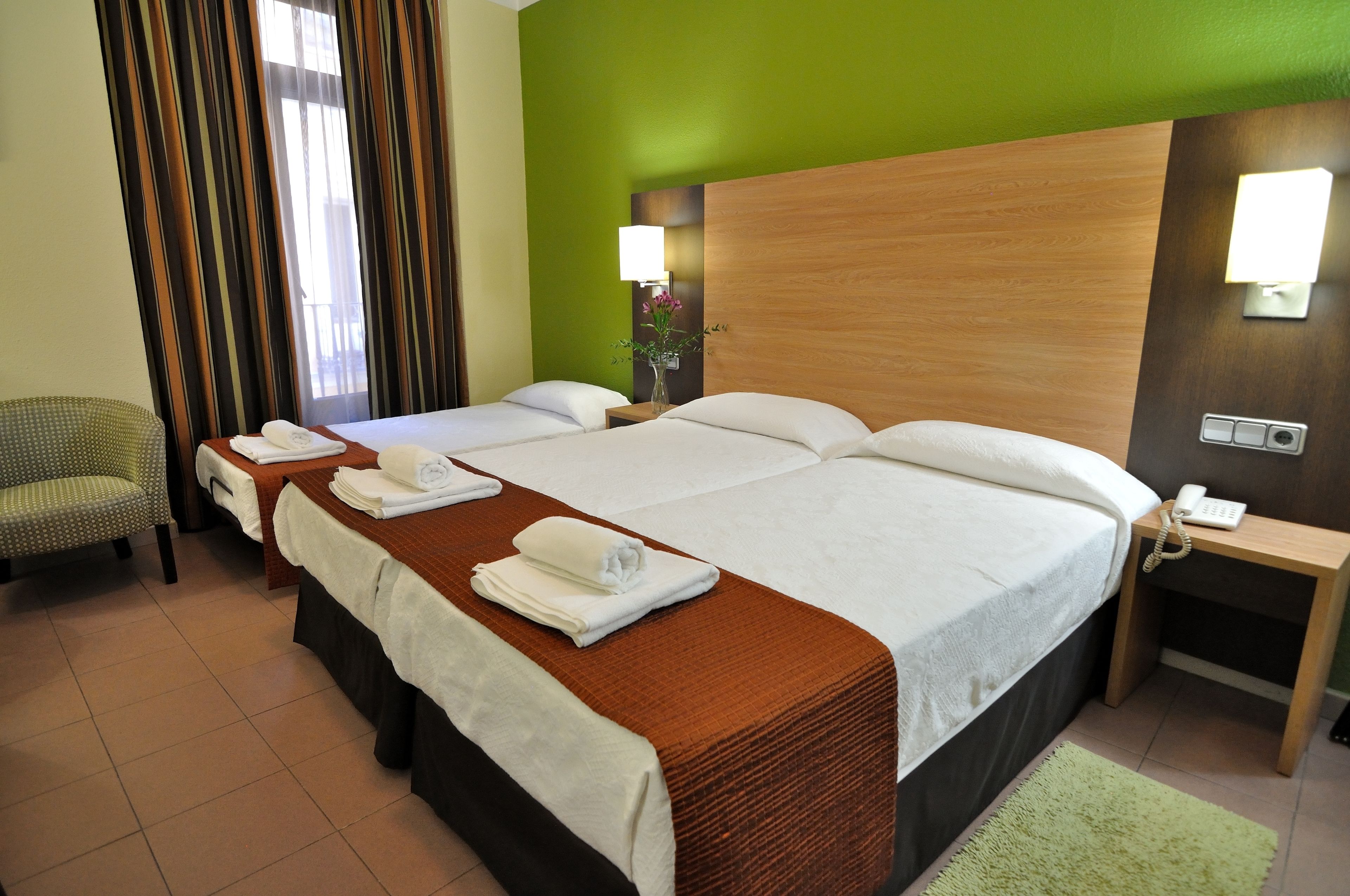 Habitación de hotel con dos camas, una pared verde, cabecera de madera, una mesita de noche con lámpara y una silla.