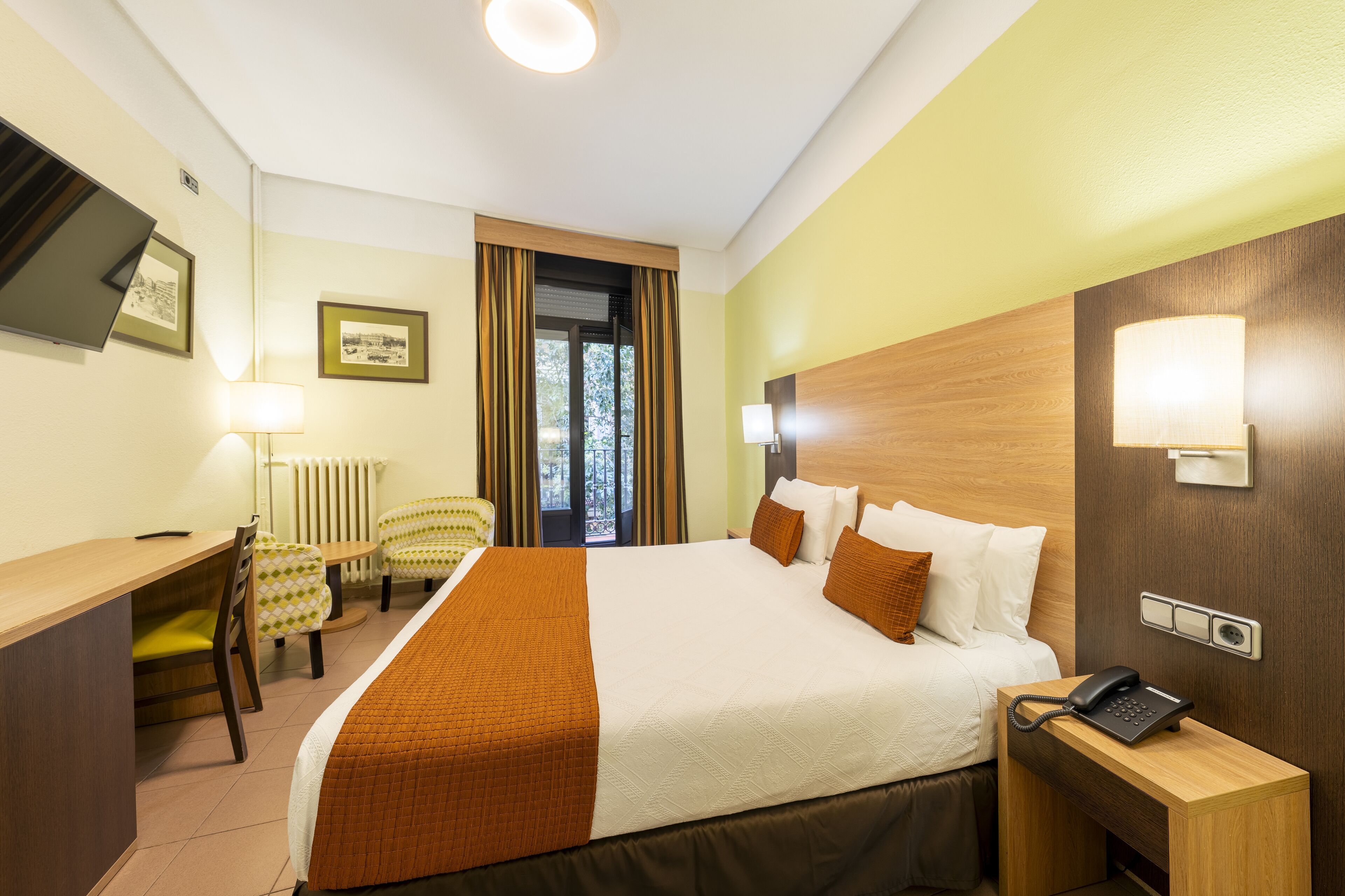 Habitación de hotel con cama, mesita de noche con teléfono, escritorio con silla, lámpara y ventana con cortinas.