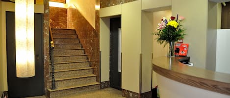 Zona de recepción de un hotel con una escalera de mármol, mostrador de recepción y un cartel en la pared.