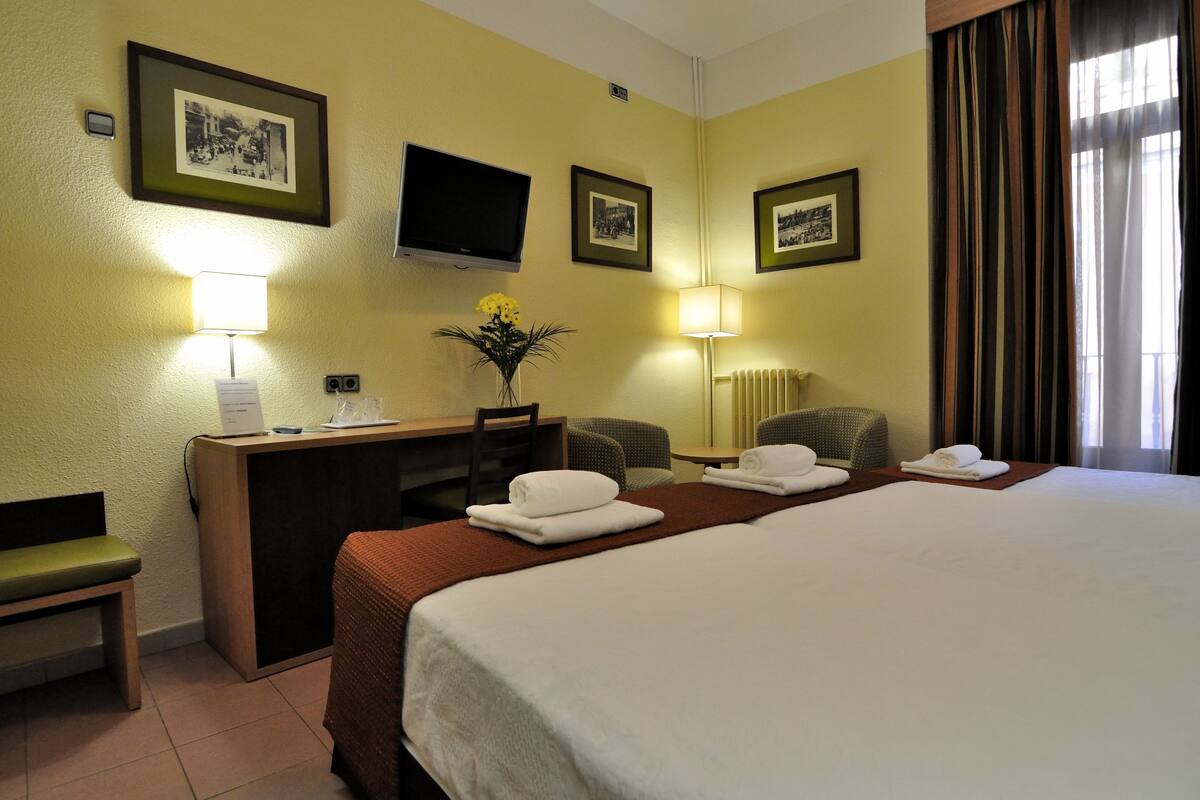 A hotel room with a bed, a desk, a chair, a TV, and framed pictures on the wall.