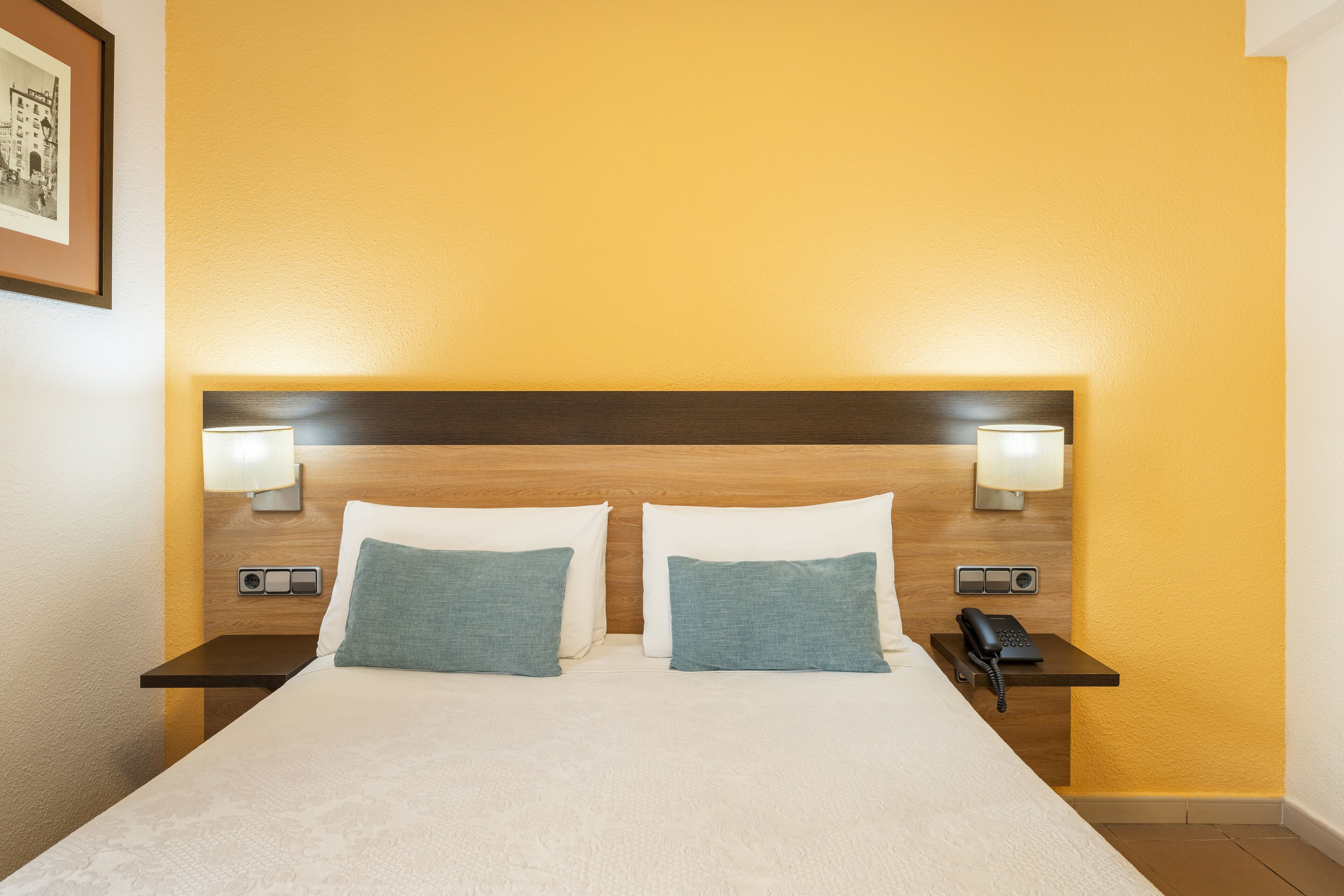 A hotel room with a large bed, two pillows, bedside lamps, a nightstand with a phone, and a framed picture on the wall.