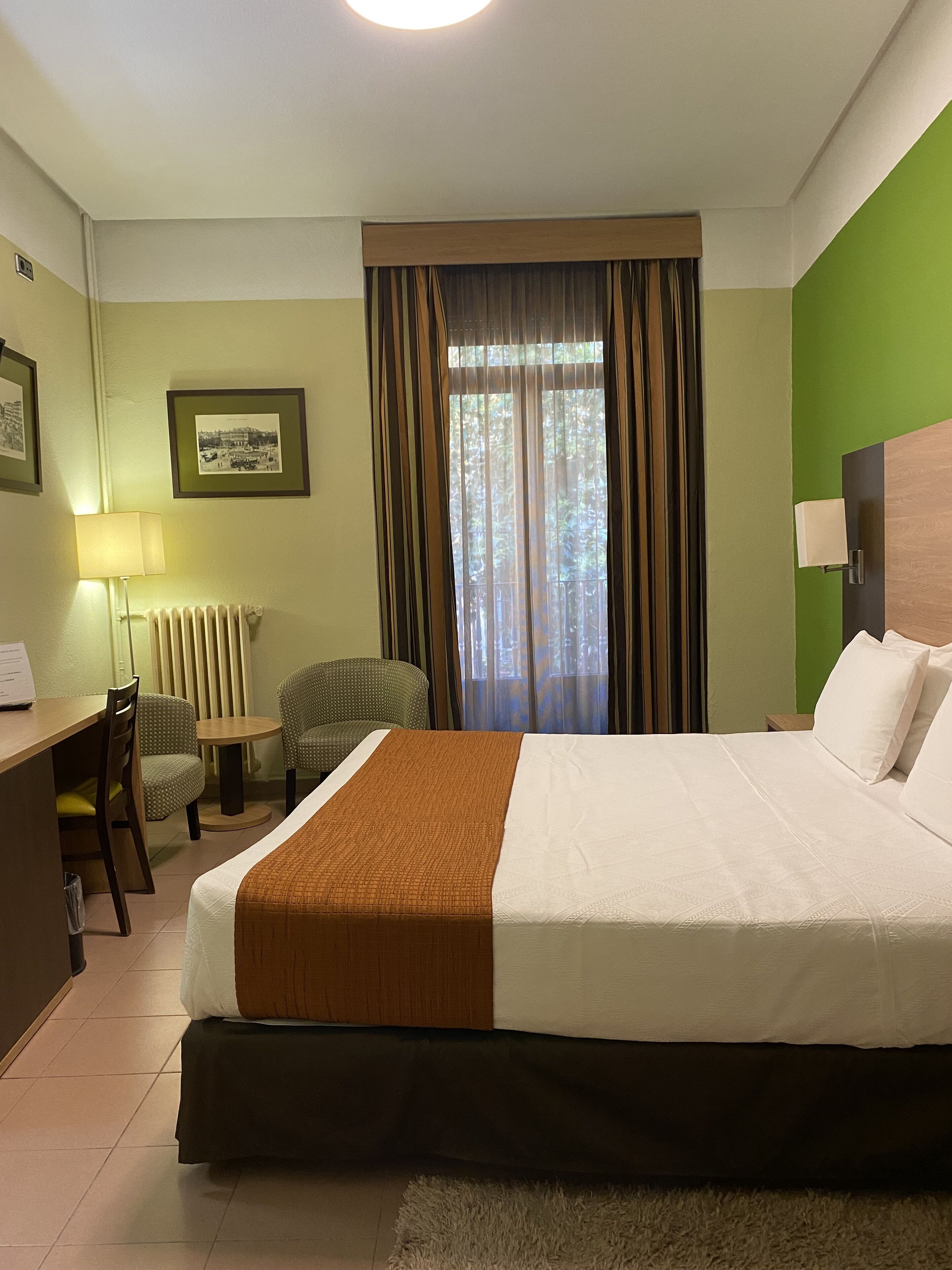 A hotel room with a bed, a desk, two chairs, a lamp, a picture on the wall, and a window with curtains.