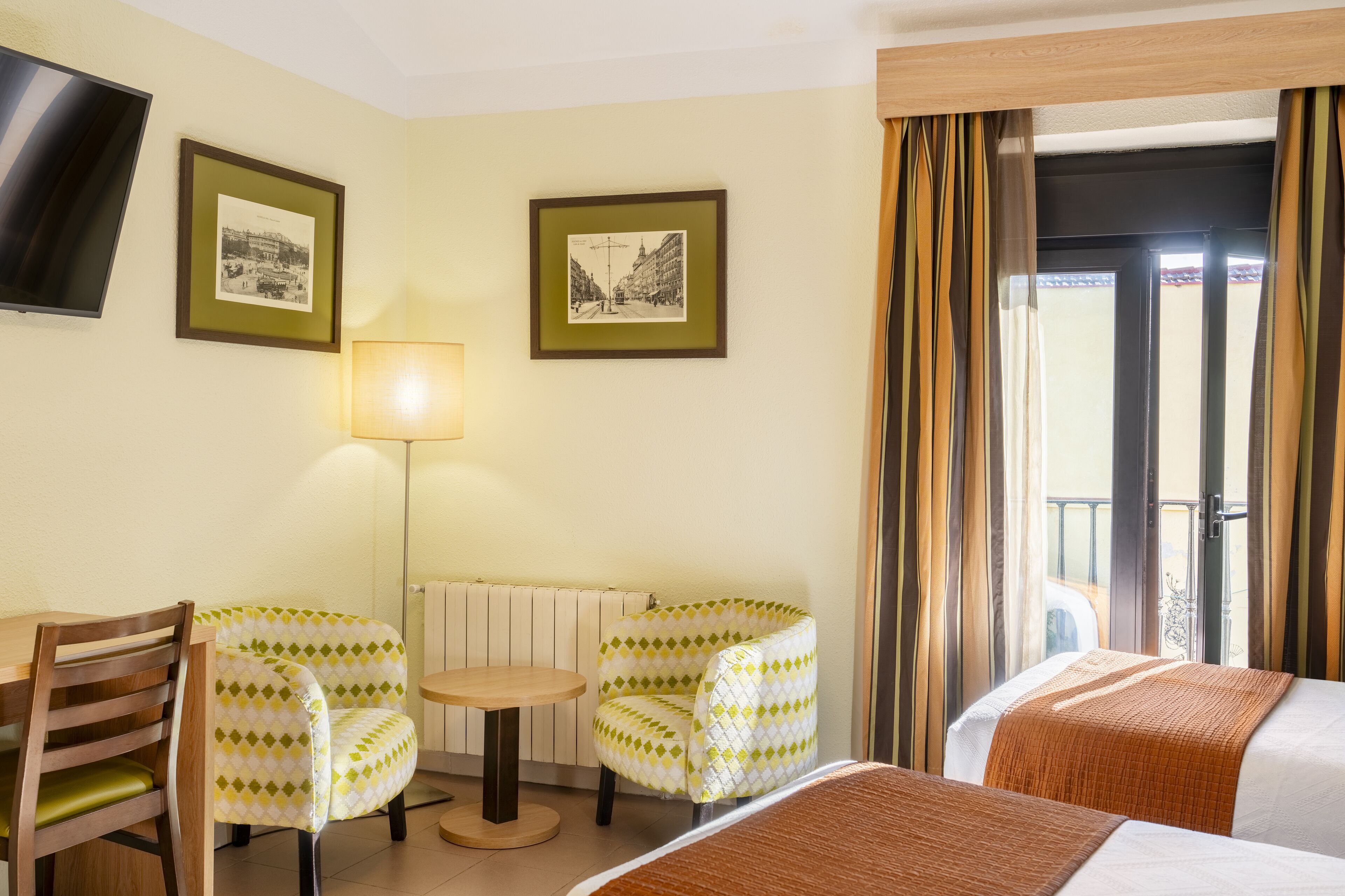 A hotel room with two chairs, a small table, a bed, a TV mounted on the wall, and two framed pictures.