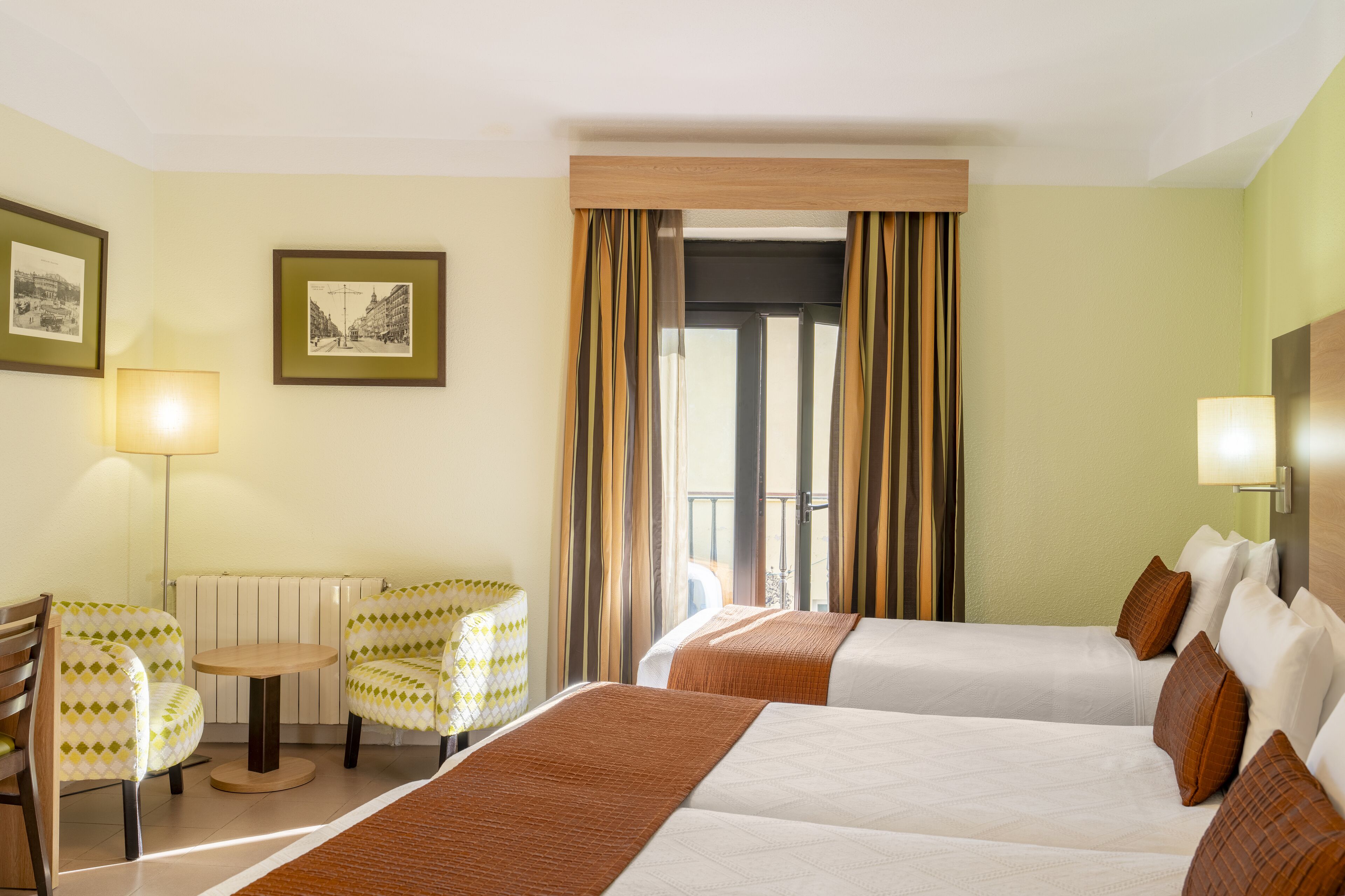 A hotel room with two beds, a small table, two chairs, a lamp, and framed pictures on the wall.