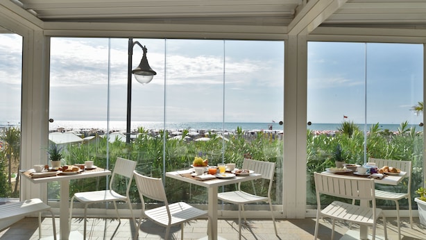 Un patio cubierto con muebles blancos y una mesa preparada para el desayuno, con vista a una playa con sombrillas y sillas de descanso.