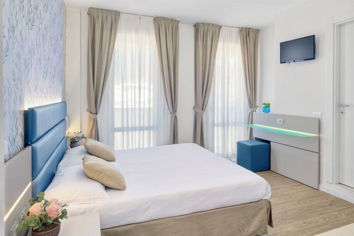 Un dormitorio moderno con cama, cortinas, televisor y mesita de noche.