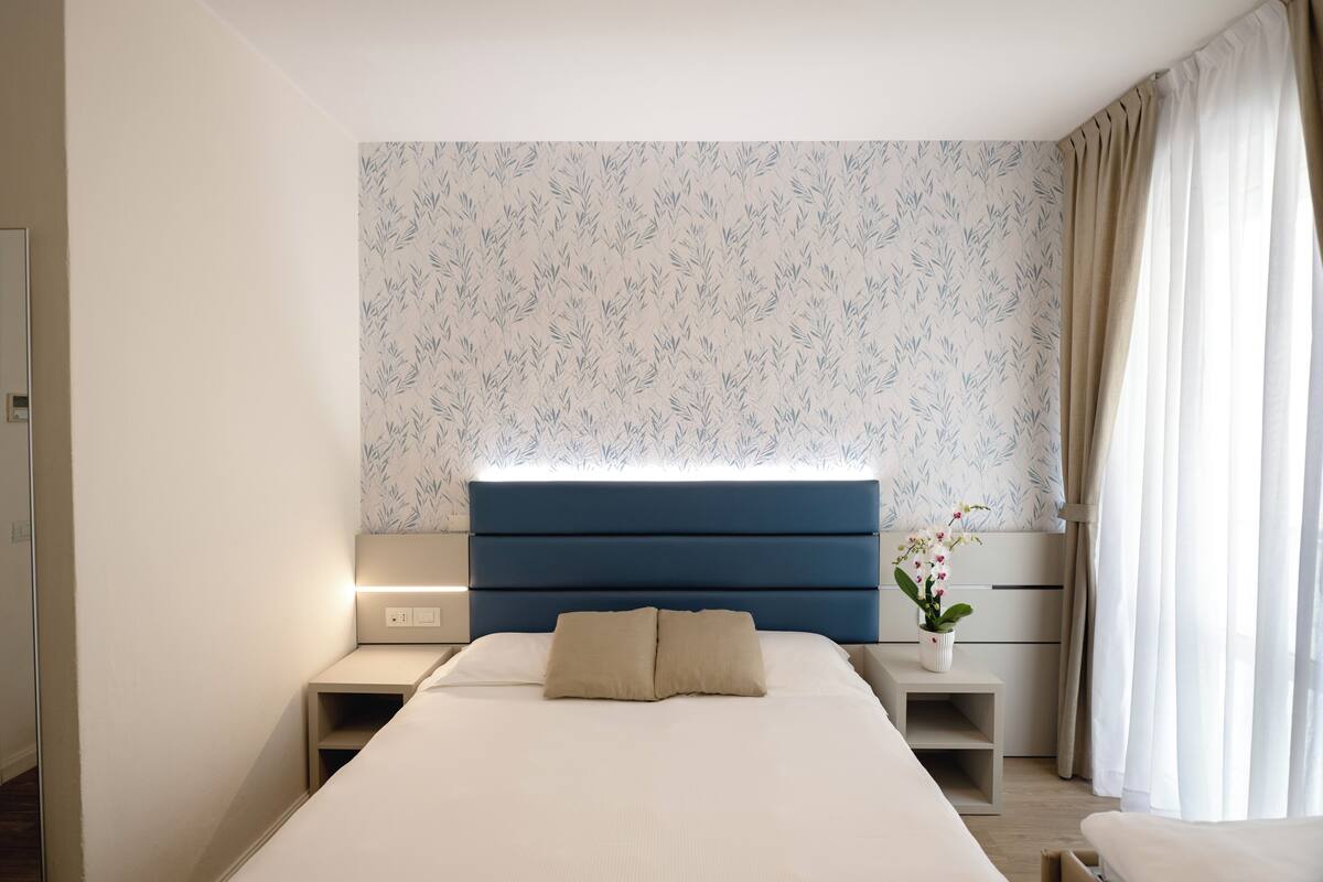Un dormitorio moderno con una cama grande, una mesita de noche con lámpara, un aparador y una ventana con cortinas.