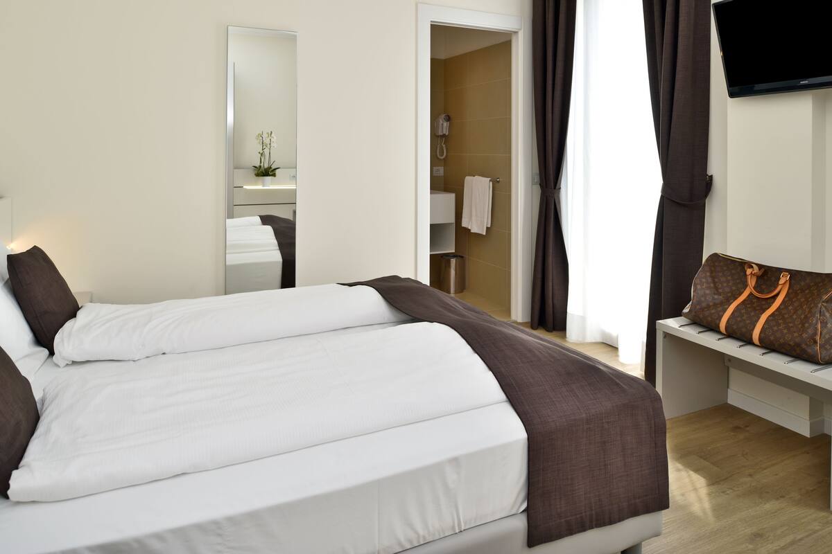 Habitación de hotel con una cama, un banco con un bolso, un televisor y un baño visible a través de una puerta abierta.