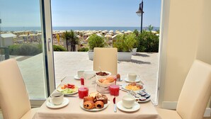 Una mesa de desayuno con diversas opciones de comida y bebidas, con vista a una playa con sombrillas y palmeras.