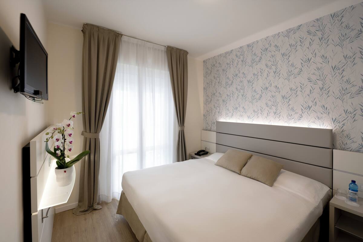Una habitación de hotel moderna con una cama grande, una mesita de noche, un televisor de pantalla plana montado en la pared, una orquídea en maceta y una ventana con cortinas.