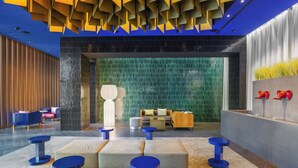 A modern lobby with a seating area featuring blue stools and cushioned benches, a reception desk, and a decorative wall with red objects. - Hotel Glam Milano (Milan)