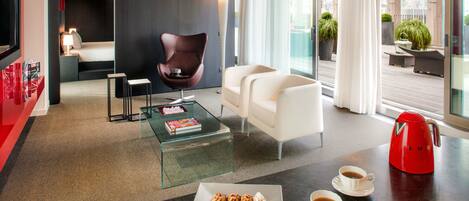 A modern hotel room with a glass coffee table, a sofa, a chair, a dining table with pastries, and a red kettle.