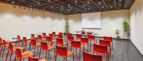 A conference room with red chairs and a projector screen displaying "GLAM Hotel".