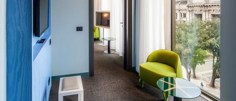 A hotel room with a television mounted on a blue wall, a green armchair, a small white table, and a view of a historic building through a large window.
