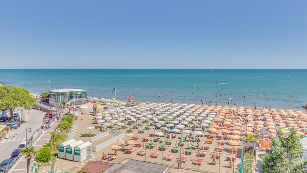 A beach with numerous umbrellas and sun loungers, a clear blue sky, and the sea in the background. - Hotel Austria (Caorle)