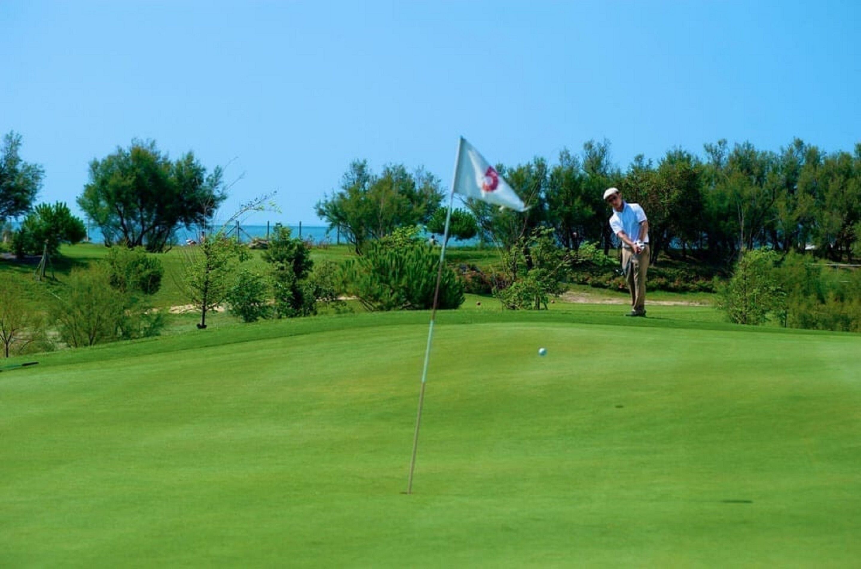 A golf course with a flag, a flagpole, and a golfer in a white shirt and khaki pants.