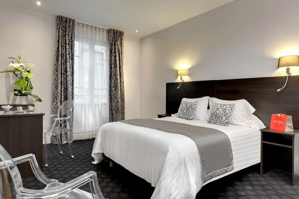 A hotel room with a large bed, two bedside lamps, a desk with a lamp, a chair, and a potted plant.