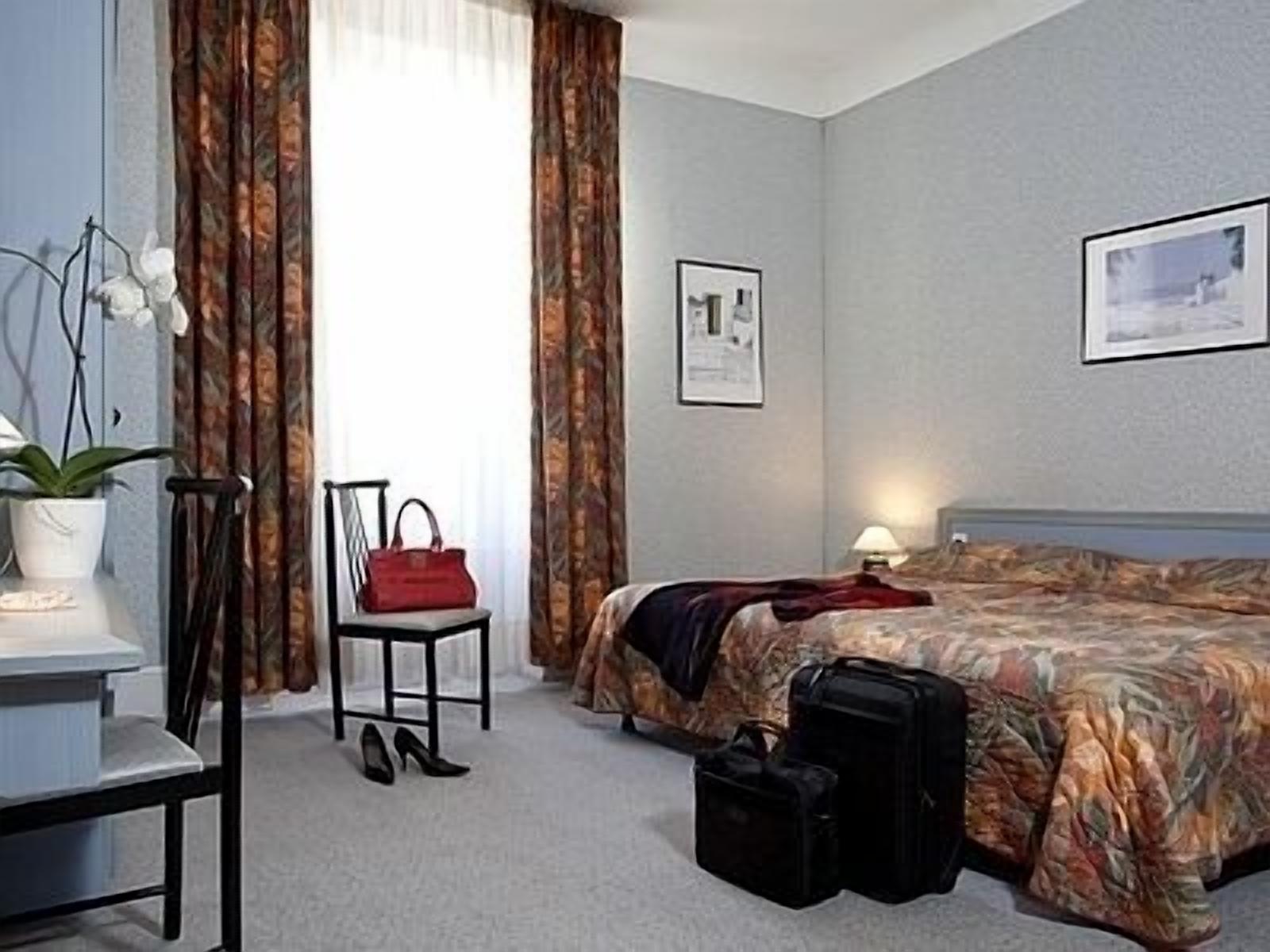 A hotel room with a bed, a chair, a desk, a lamp, and a suitcase.